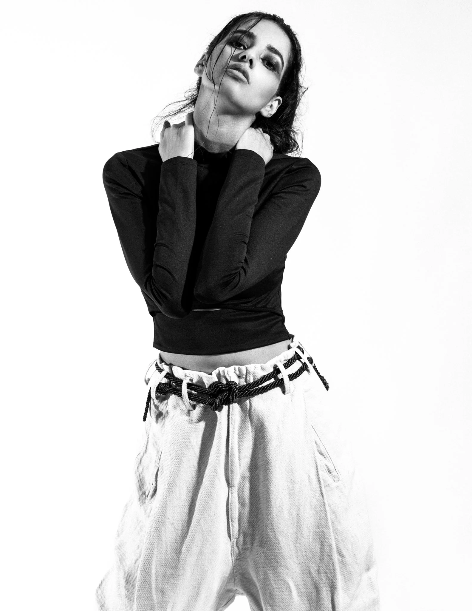 Black and white photograph of a woman with dark hair pulled back, wearing a long-sleeve black shirt and light-colored wide-leg pants with a rope belt, posing against a plain white background.