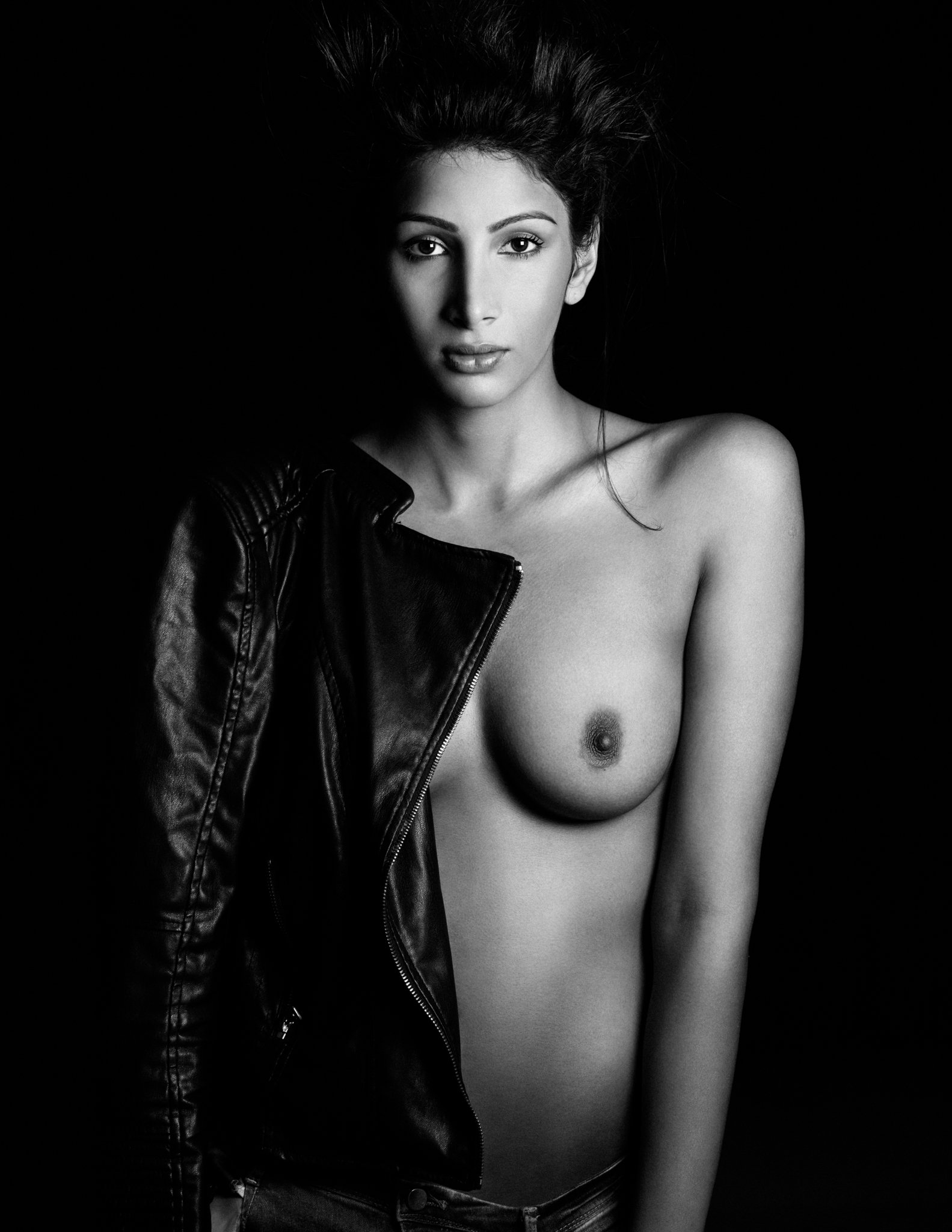 Black and white portrait of a young woman with dark hair, wearing a leather jacket, and showing her bare shoulder and breast, against a black background.