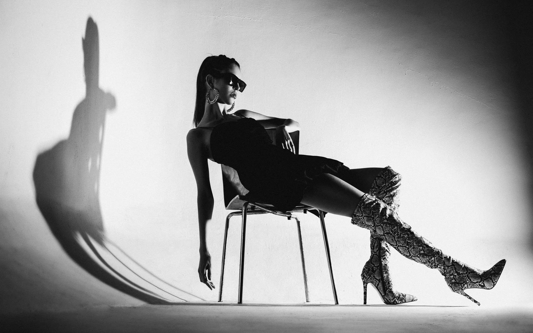 A woman sitting on a chair wearing sunglasses, hoop earrings, a strapless dress, and high-heeled snakeskin boots, with her shadow cast onto the white background.