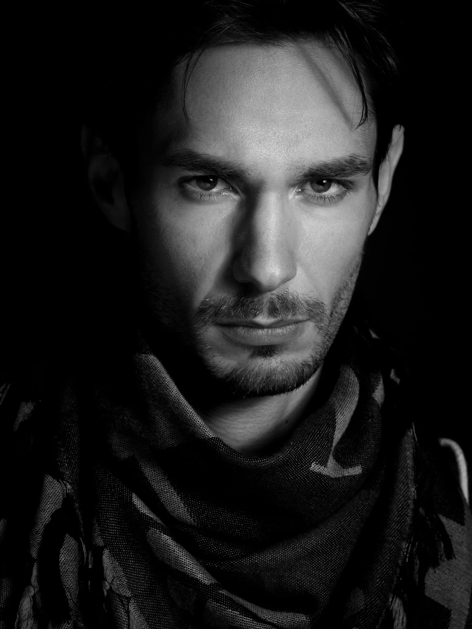 Black and white close-up portrait of a man with dark hair and a serious expression, wearing a camouflage scarf.