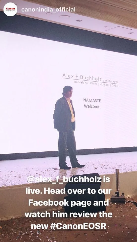 A man standing on a stage with a large screen behind him displaying his name, 'Alex F Buchholz', and the words 'NAMASTE Welcome'. The text on the screen indicates the event is in multiple cities including Barcelona, Delhi, Mumbai, and Dubai. The phot