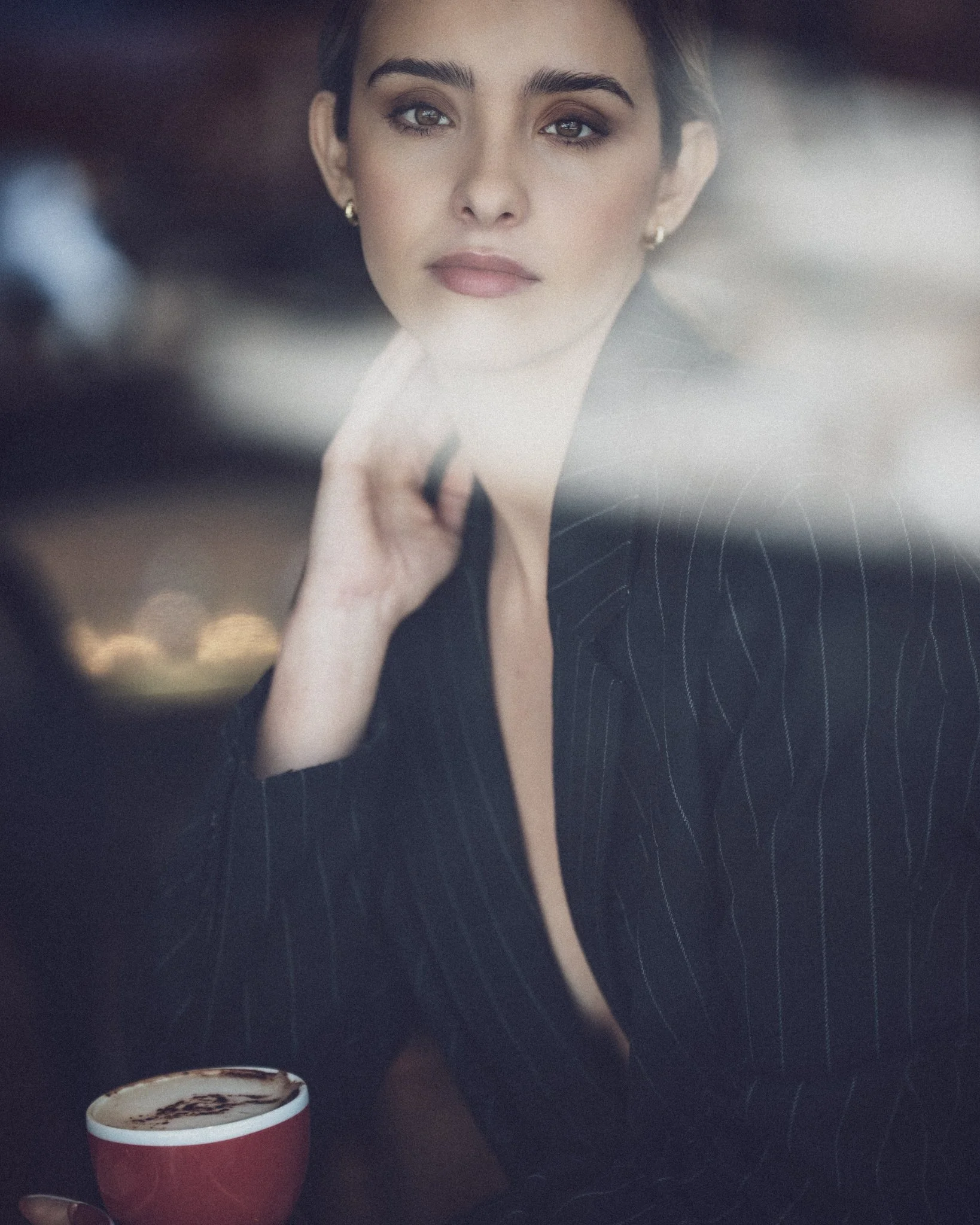 A woman with dark hair and earrings sitting at a table with a red cup of hot chocolate topped with whipped cream and chocolate shavings.