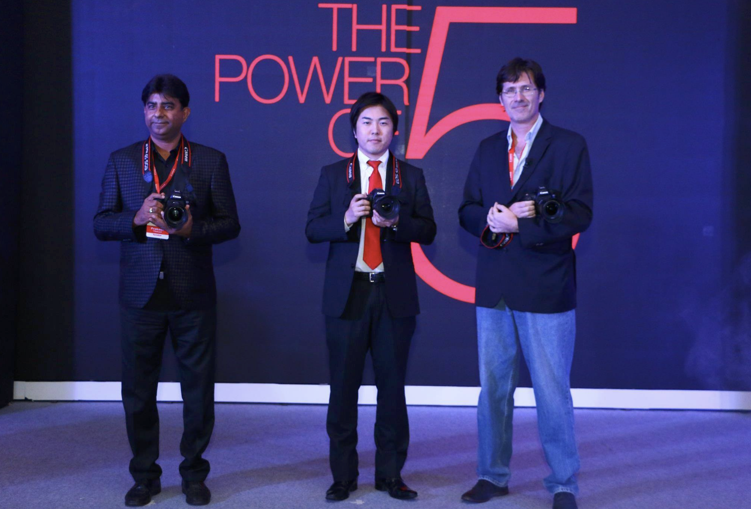 Three men in business attire holding cameras at a conference or event with a dark backdrop displaying the text 'The Power of 5' in red.
