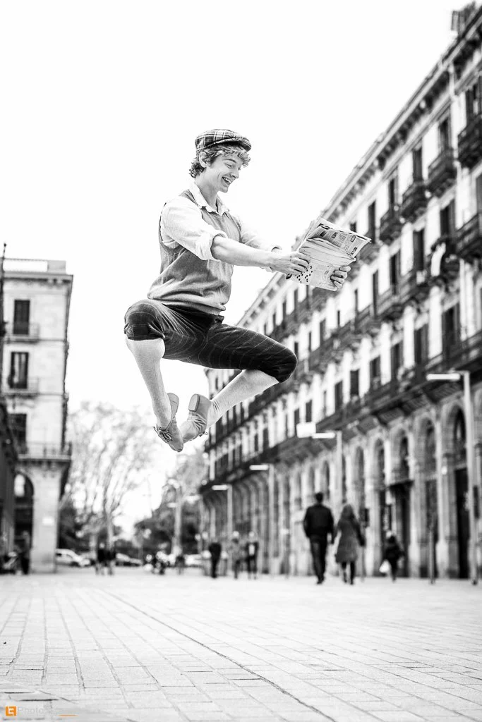 A woman jumping in the air while holding a magazine or newspaper in an urban street with old buildings and pedestrians.