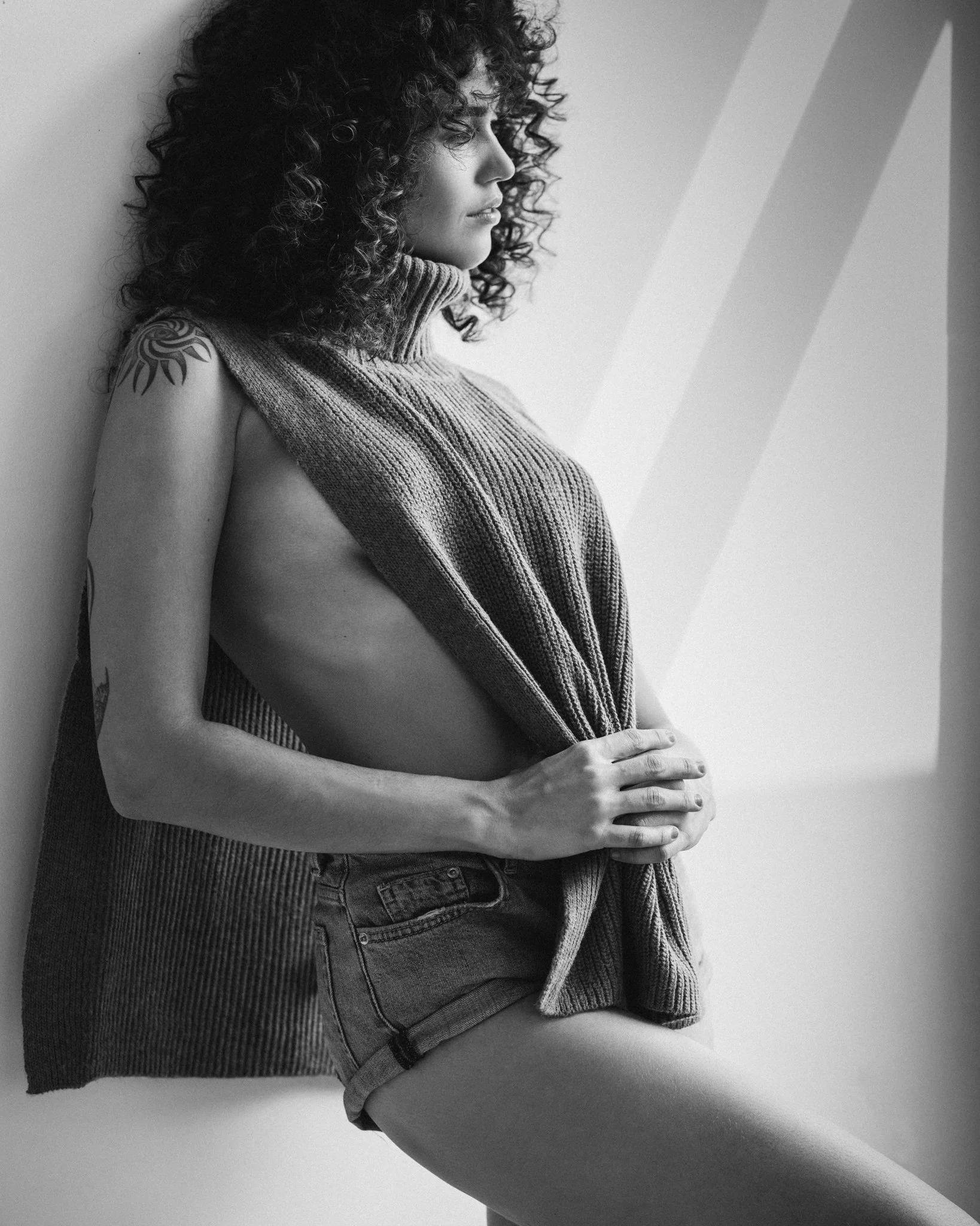 A black-and-white photograph of a young woman with curly hair, wearing a sleeveless turtleneck sweater and denim shorts, sitting against a wall with geometric patterns.