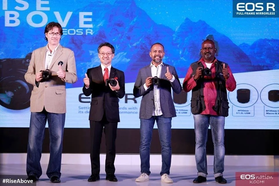 Four men including Alex F Buchholz standing on stage holding cameras, posing for a photo at a camera launch event with a blue background and the text 'Rise Above' and 'EOS R'.