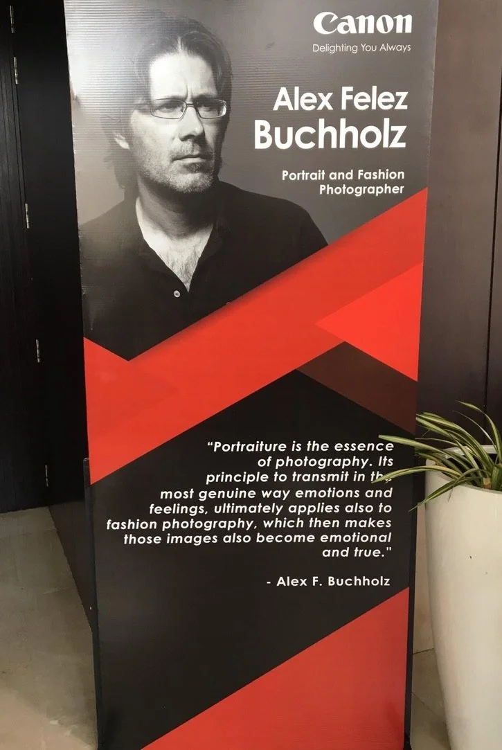 A tall vertical poster displays a black-and-white photograph of a man with glasses and dark hair, dressed in a dark shirt. The poster features the Canon logo and promotional text about Alex Felez Buchholz, a portrait and fashion photographer. The bac