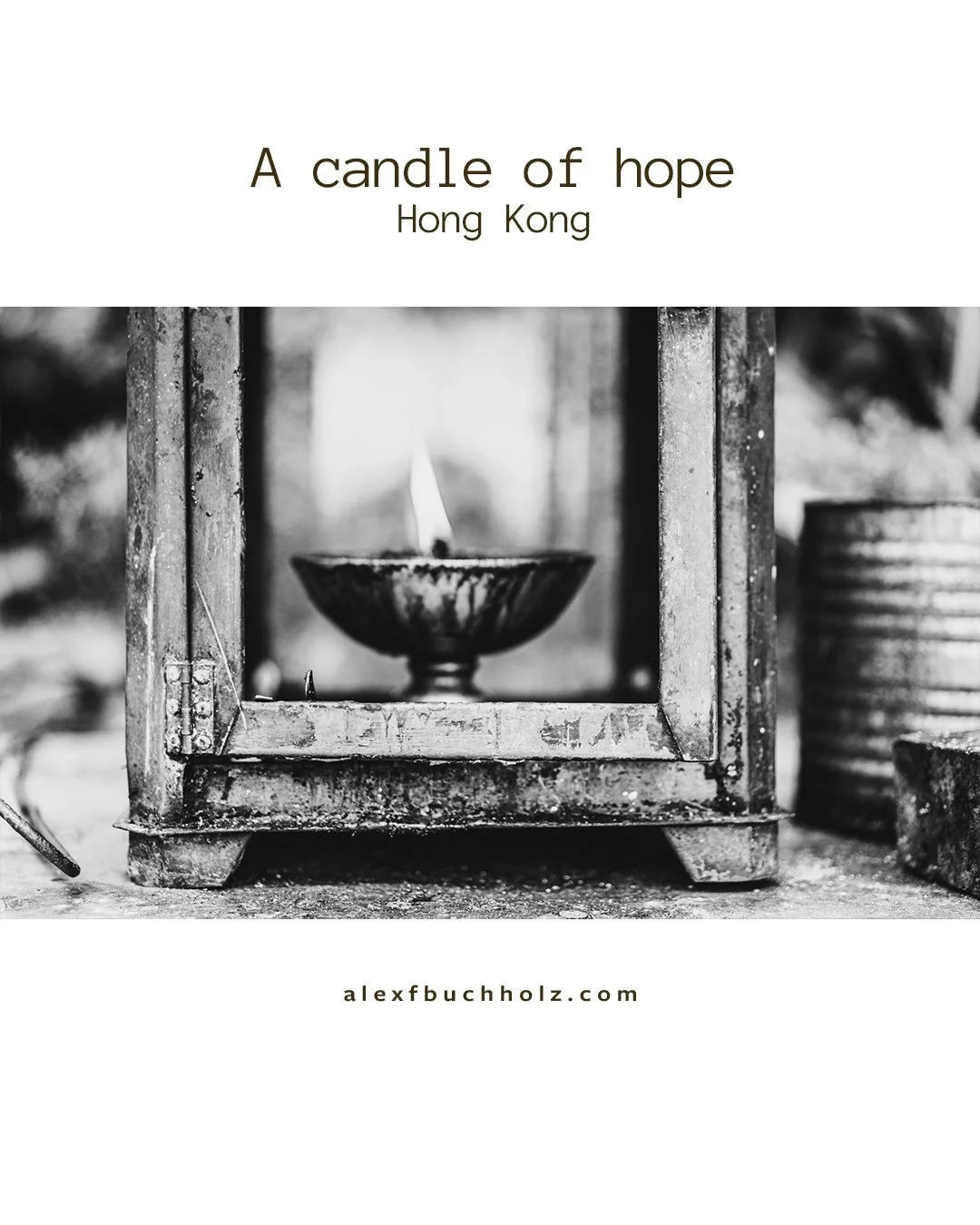 Black and white photo of a candle in a metal holder inside a glass enclosure, with the flame lit, situated outdoors next to a metal container.