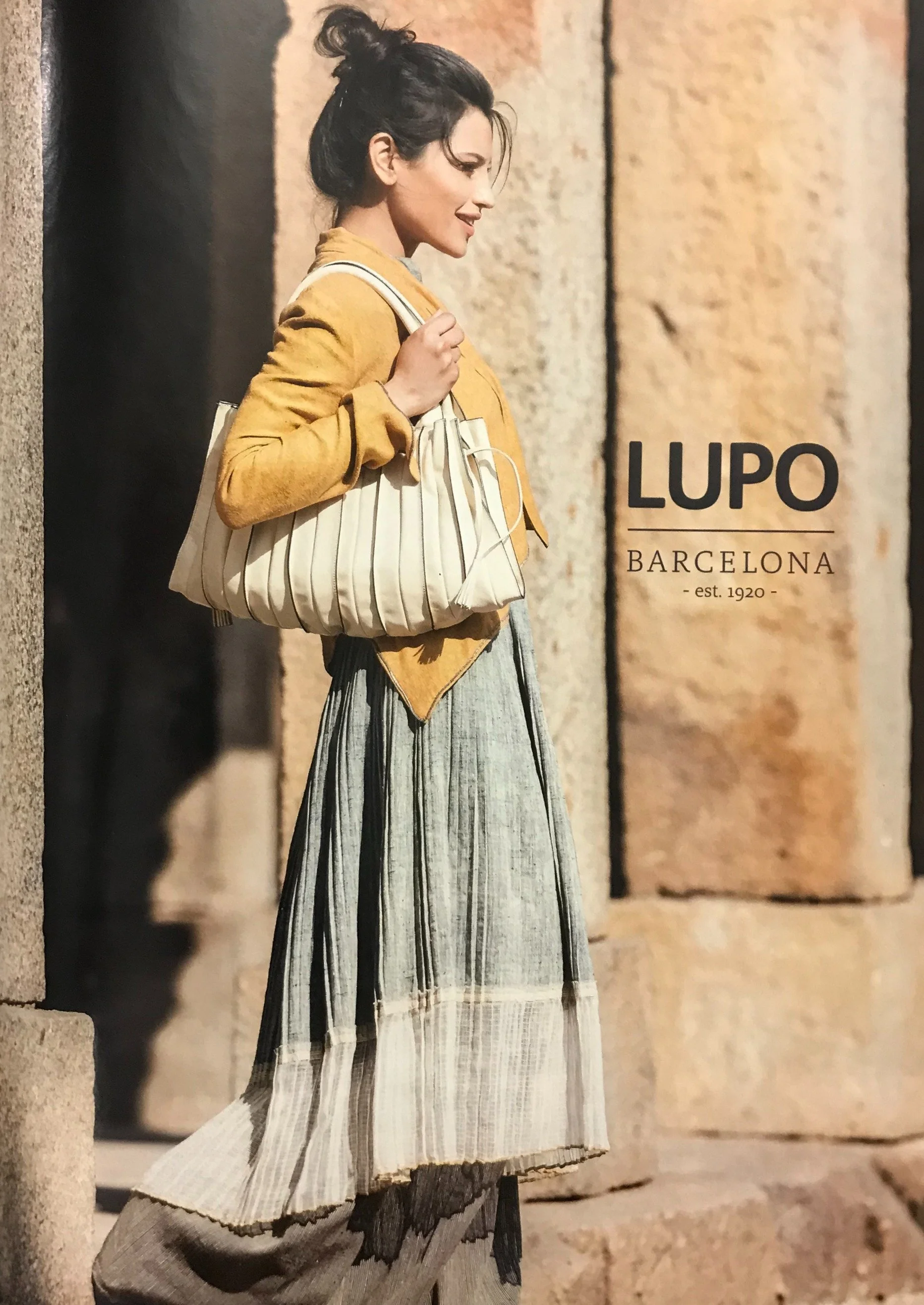A woman walking outdoors at sunset, carrying a striped beige and white bag, wearing a mustard yellow coat and a striped gray and white skirt, with a stone wall in the background and the text 'LUPO BARCELONA est. 1920'.
