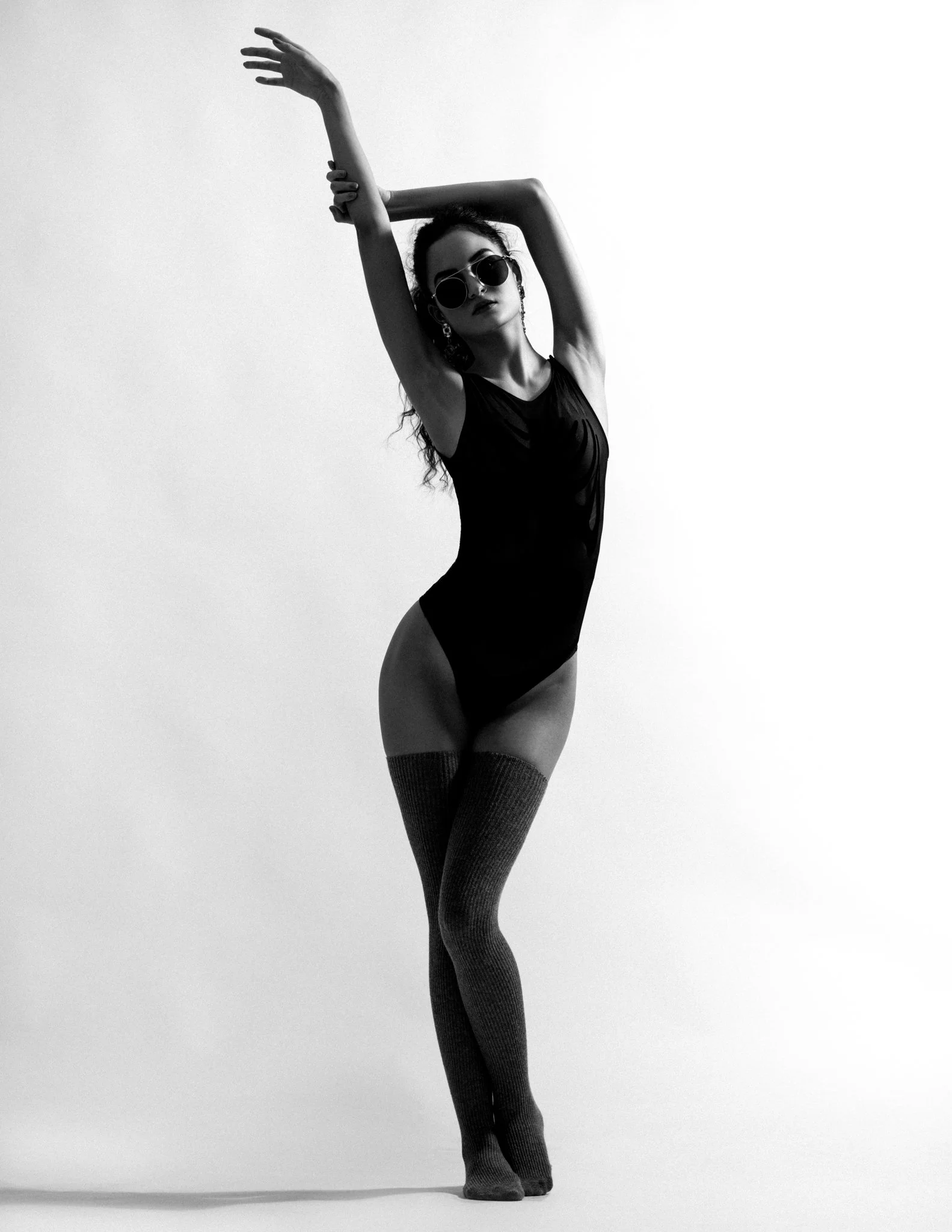 A woman in black swimsuit and thigh-high socks wearing sunglasses, posing with arms raised and one hand touching her head, in black-and-white studio photo.
