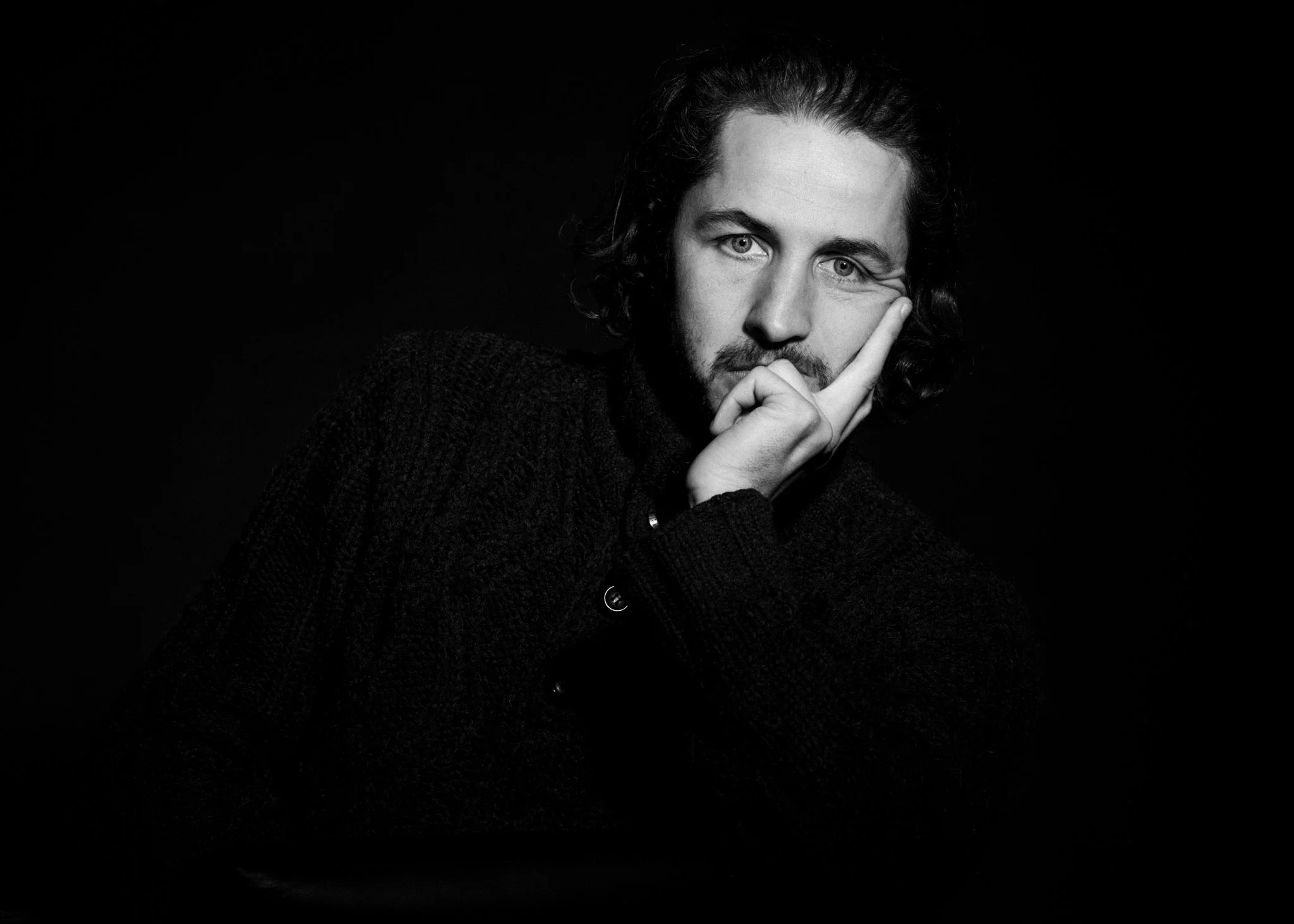 Black and white portrait of a man with curly hair and a beard, resting his face on his hand with a contemplative expression, against a dark background.