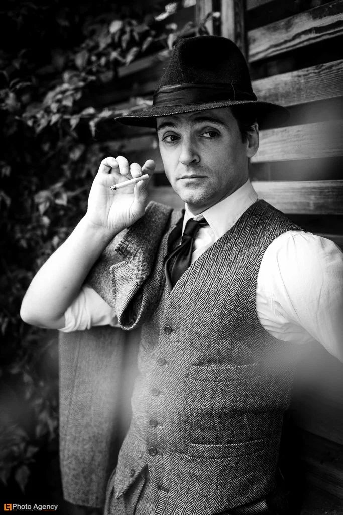 A man dressed in vintage attire with a hat, vest, and tie, holding a cigarette, standing outdoors next to a wooden fence.