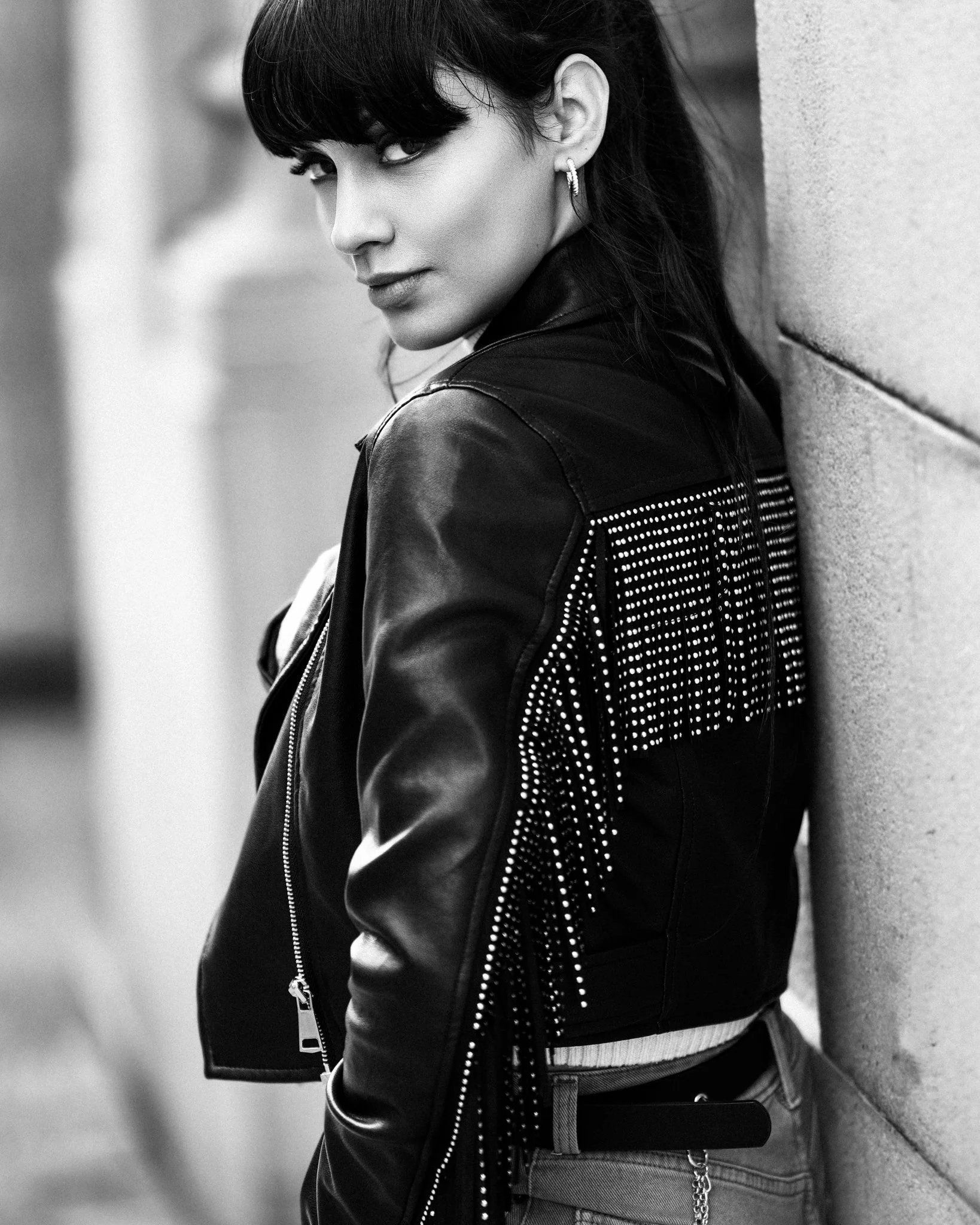 Black and white photo of a woman with dark hair and bangs, wearing a leather jacket with decorative studs and fringe, leaning against a wall and looking at the camera.