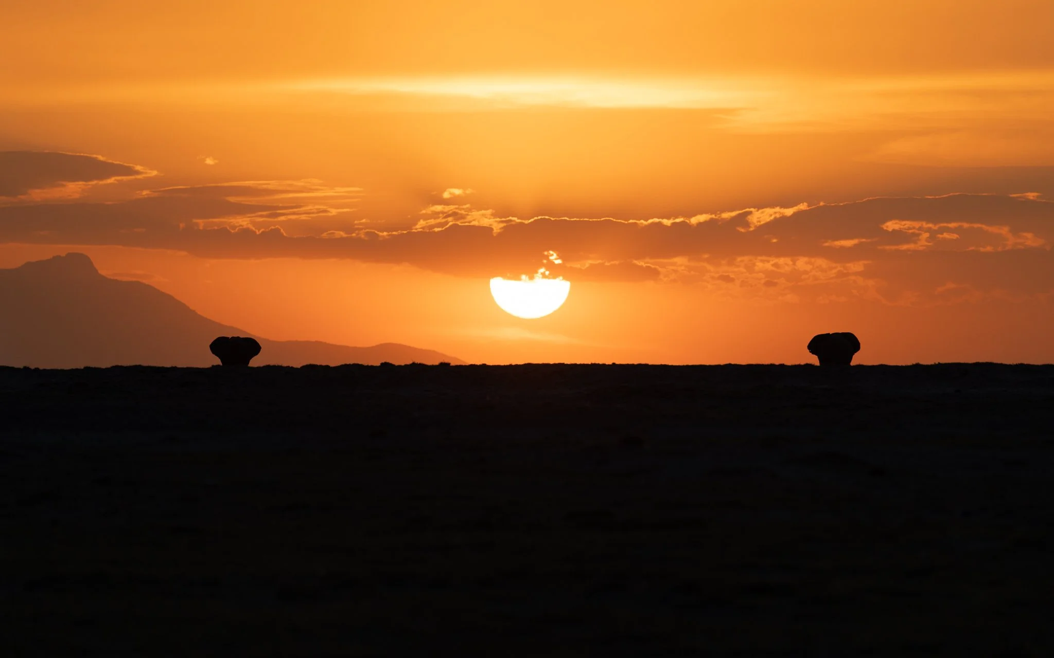 Sunset with the sun partially obscured by clouds, casting an orange glow on the sky, with two dark rocks on the horizon.
