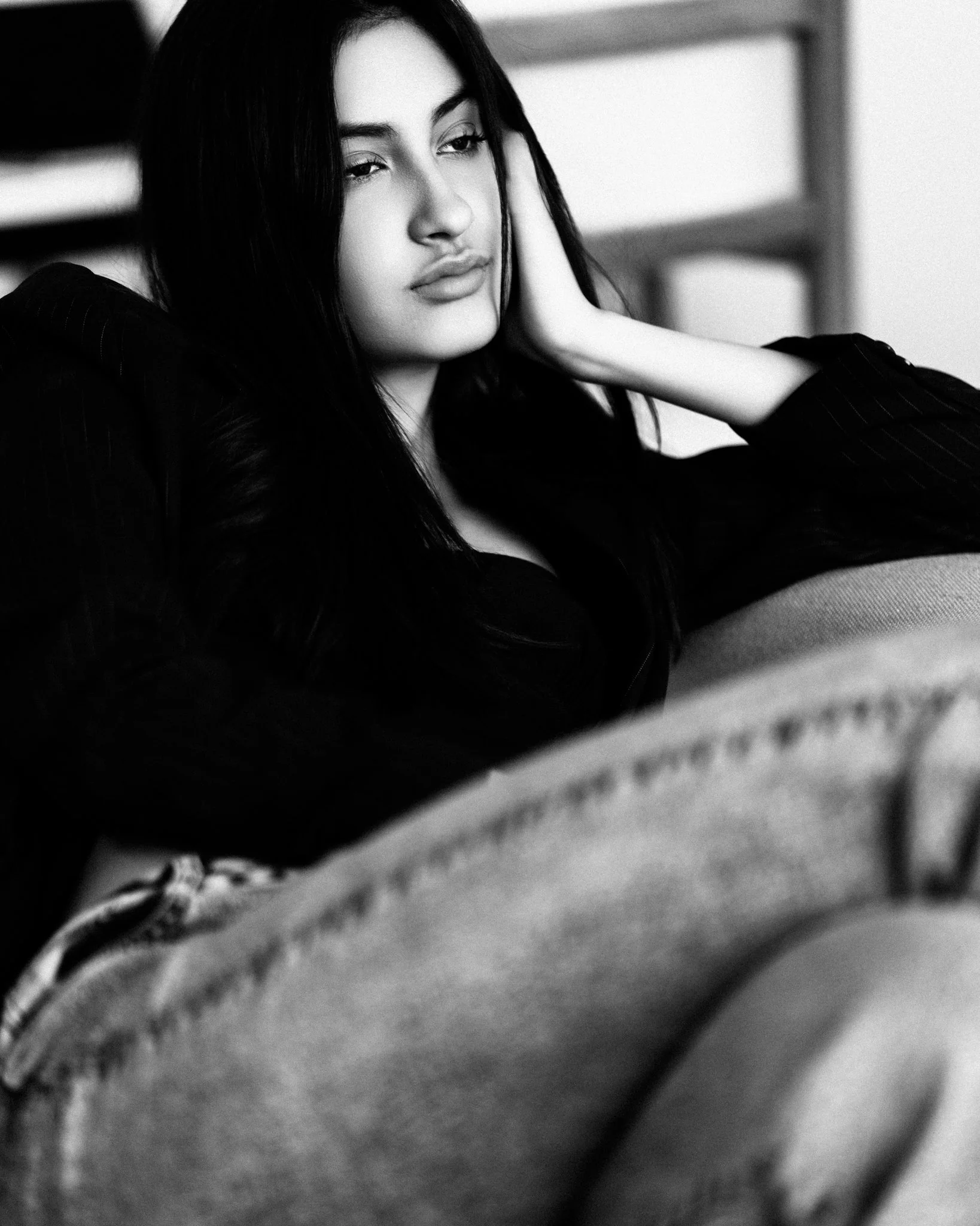 Black and white photo of a young woman with long dark hair, lying on a couch with her hand resting on her head, looking contemplative.