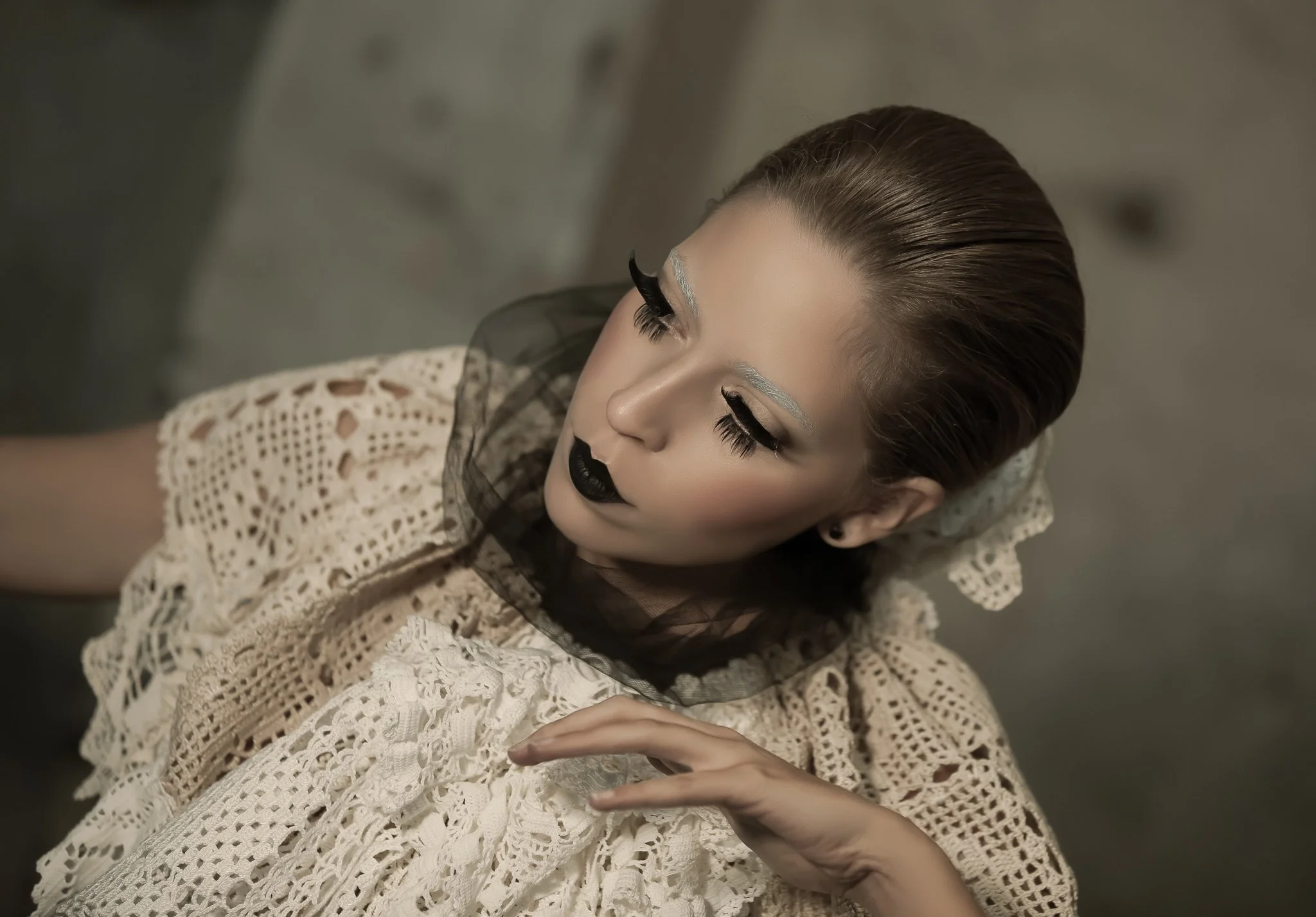 A woman with slicked-back hair and dramatic makeup, including heavy black eyeliner, dark lipstick, and metallic silver eyebrows, wearing a cream-colored crochet dress, poses with her eyes closed and hand gently touching her neck.