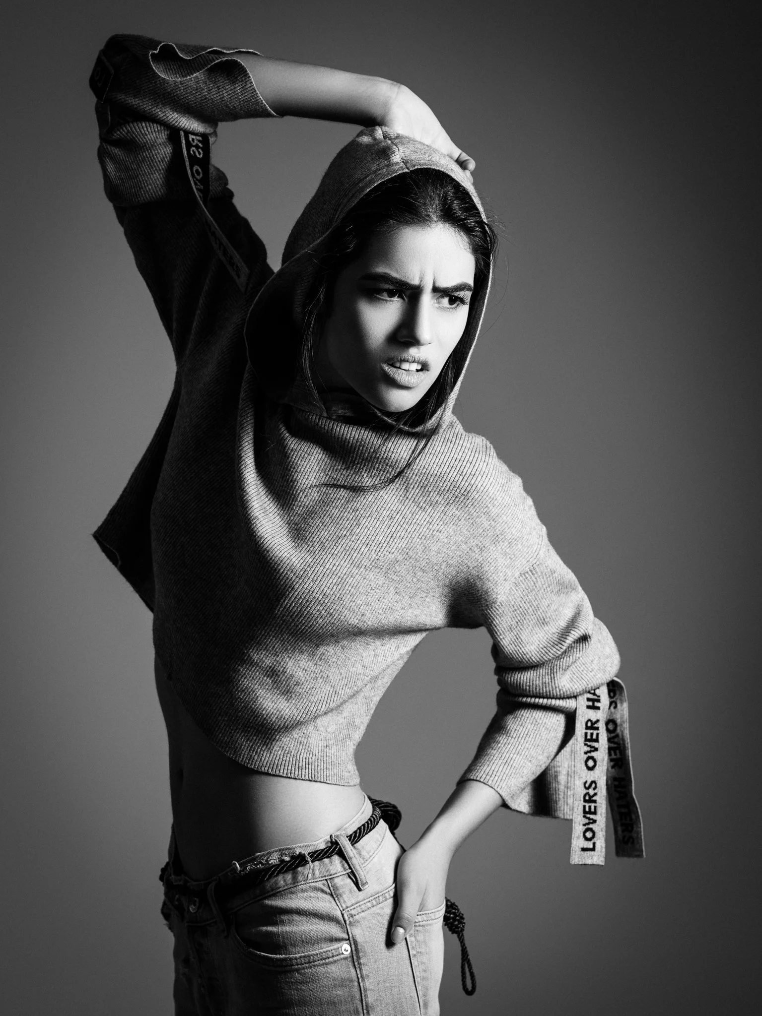 Black and white photo of a woman with a hoodie, looking confused or angry, wearing jeans with a belt, posing against a plain background.