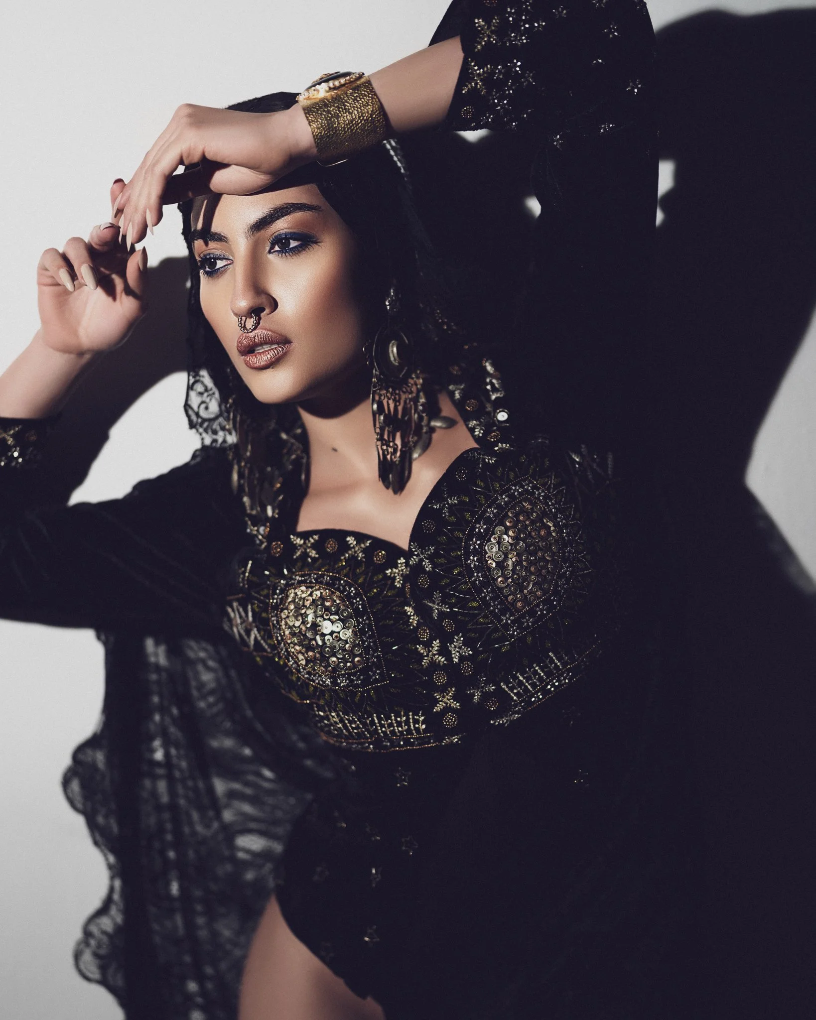 A woman wearing a black, embroidered, sequined dress with large earrings and a gold cuff bracelet. She has dark hair styled with beads, makeup, and a septum piercing. She poses with her right arm above her head and her left arm touching her forehead 