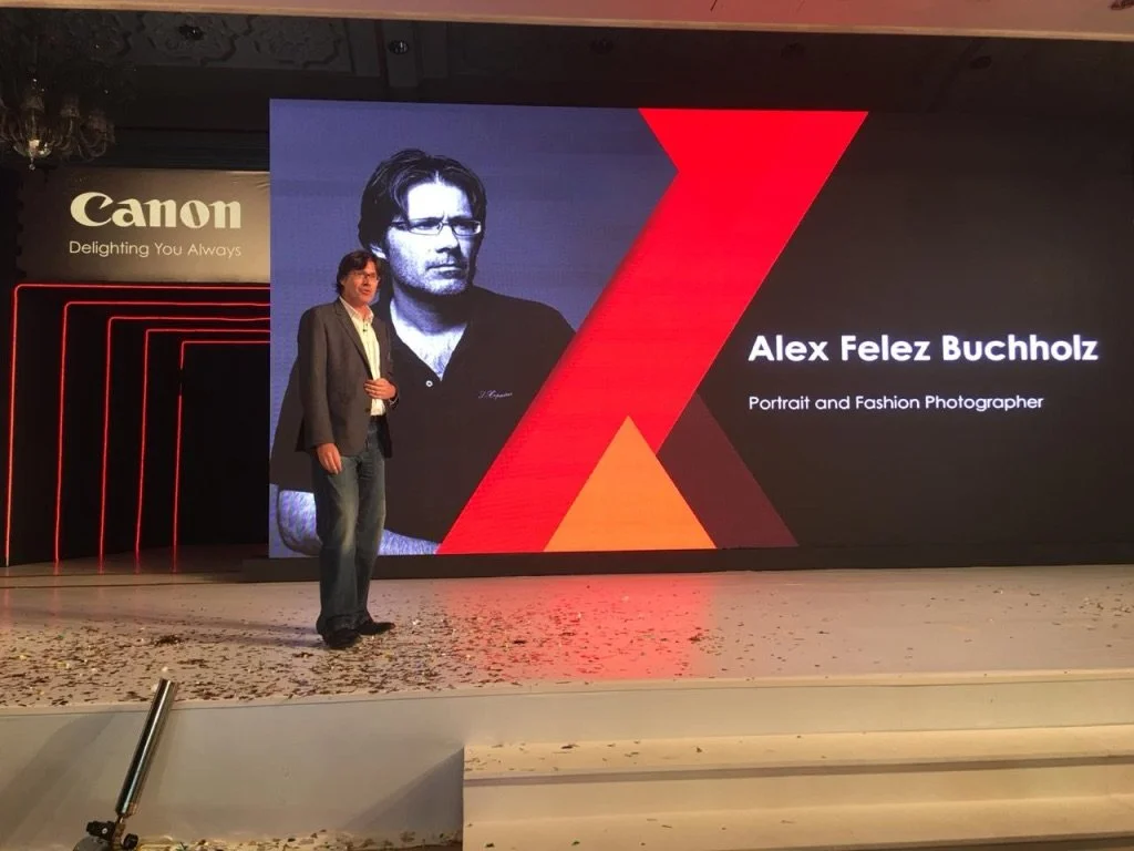 Photograph of a man standing on stage during a presentation. Behind him, a large screen displays a portrait of a man with glasses and dark hair, along with the text 'Alex Felez Buchholz, Portrait and Fashion Photographer.' To the left, there is a 'Ca