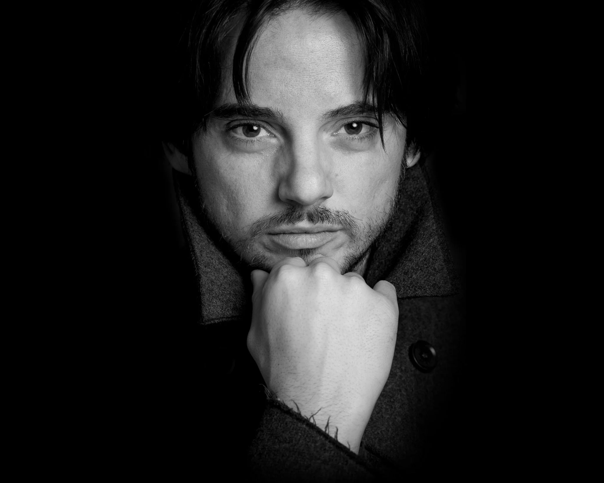 Black and white portrait of a young man with dark hair, looking directly at the camera with a contemplative expression. He rests his chin on his hand, with the background fading into black.