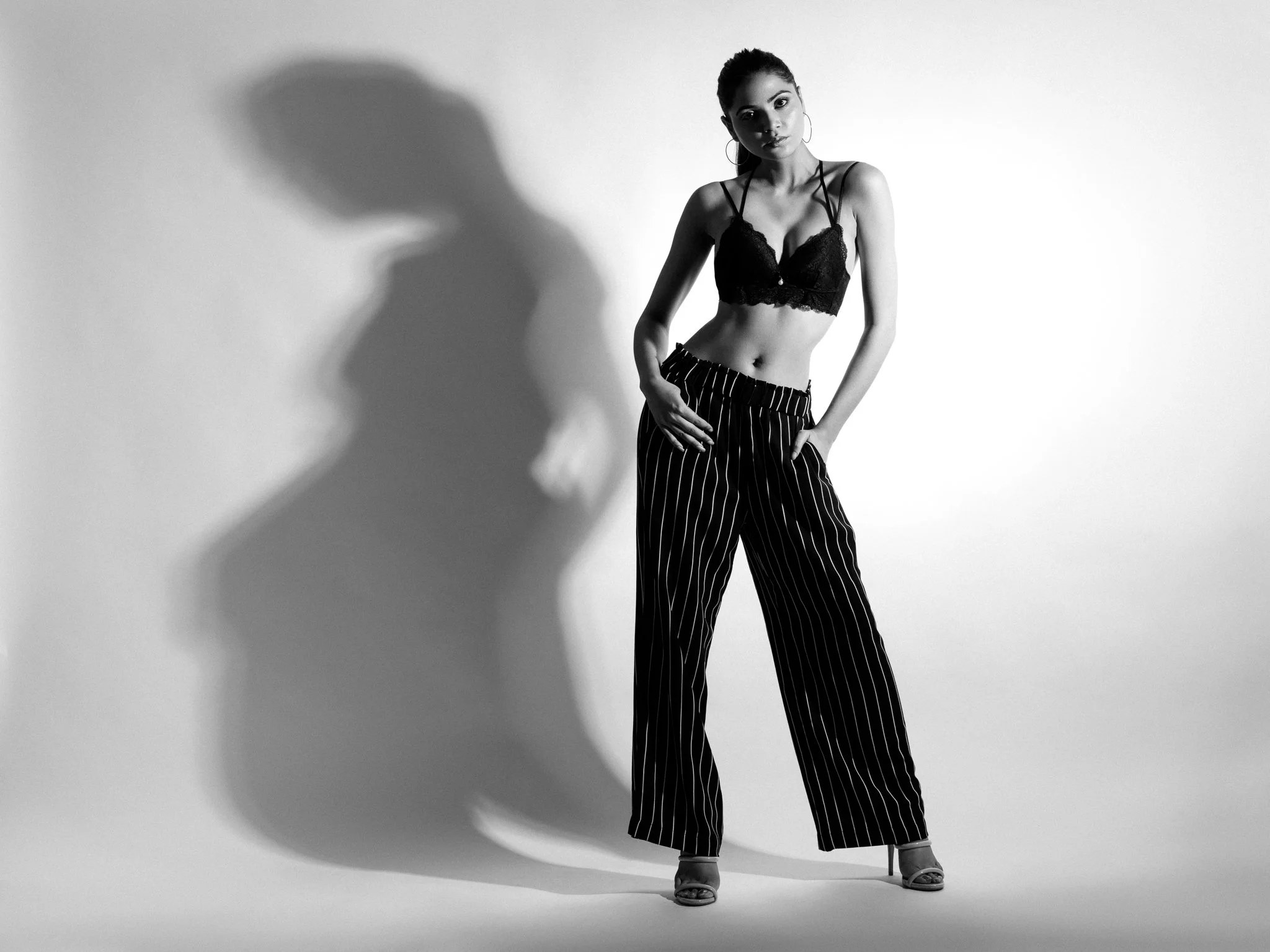 A young woman stands against a plain light-colored background, dressed in a black lace cropped top, striped wide-leg trousers, and high heels. She has hoop earrings and her hair is tied back. Her pose is relaxed, with one hand resting on her thigh an