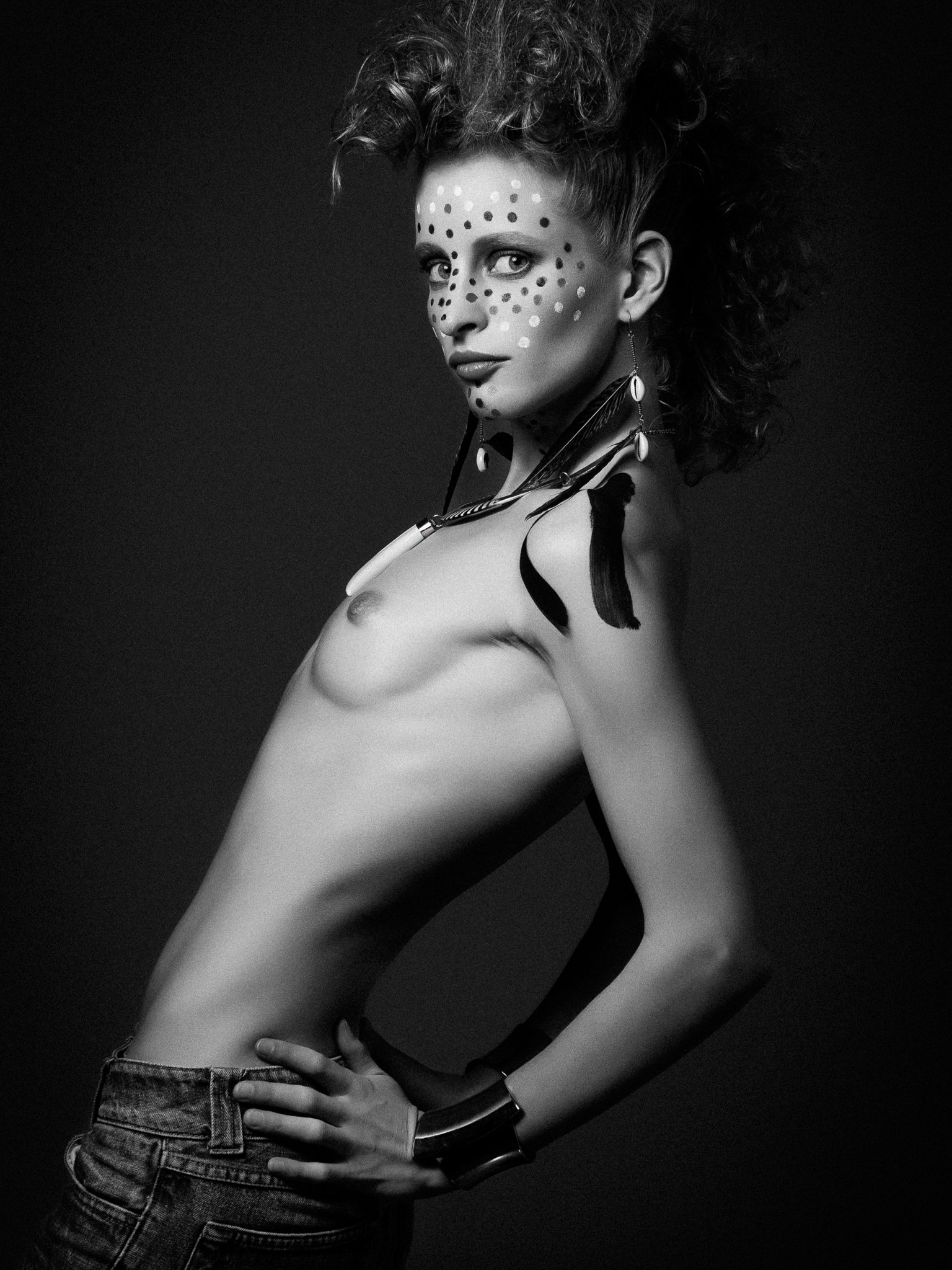 Black and white photo of a woman with curly hair and face painted with dots, wearing earrings, a necklace, and denim jeans, posing with one hand on her hip and the other on her waist, against a dark background.