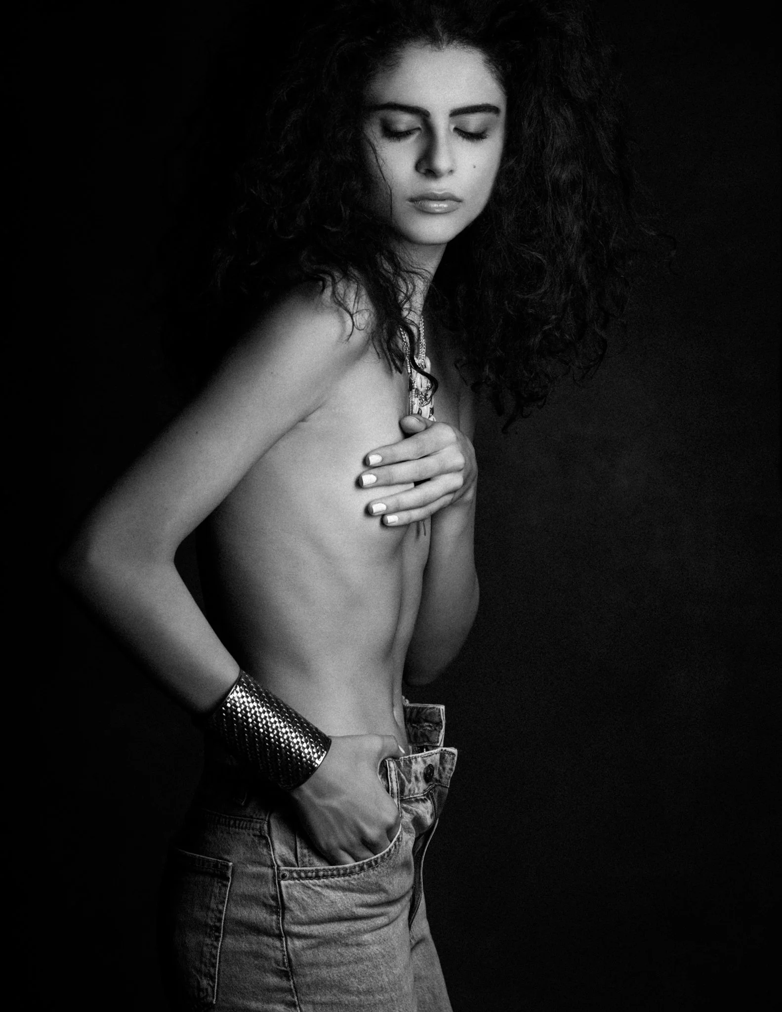 Black and white photo of a woman with curly hair, partly nude, wearing jeans, a watch, and a necklace, covering her body with her hand on her chest against a dark background.