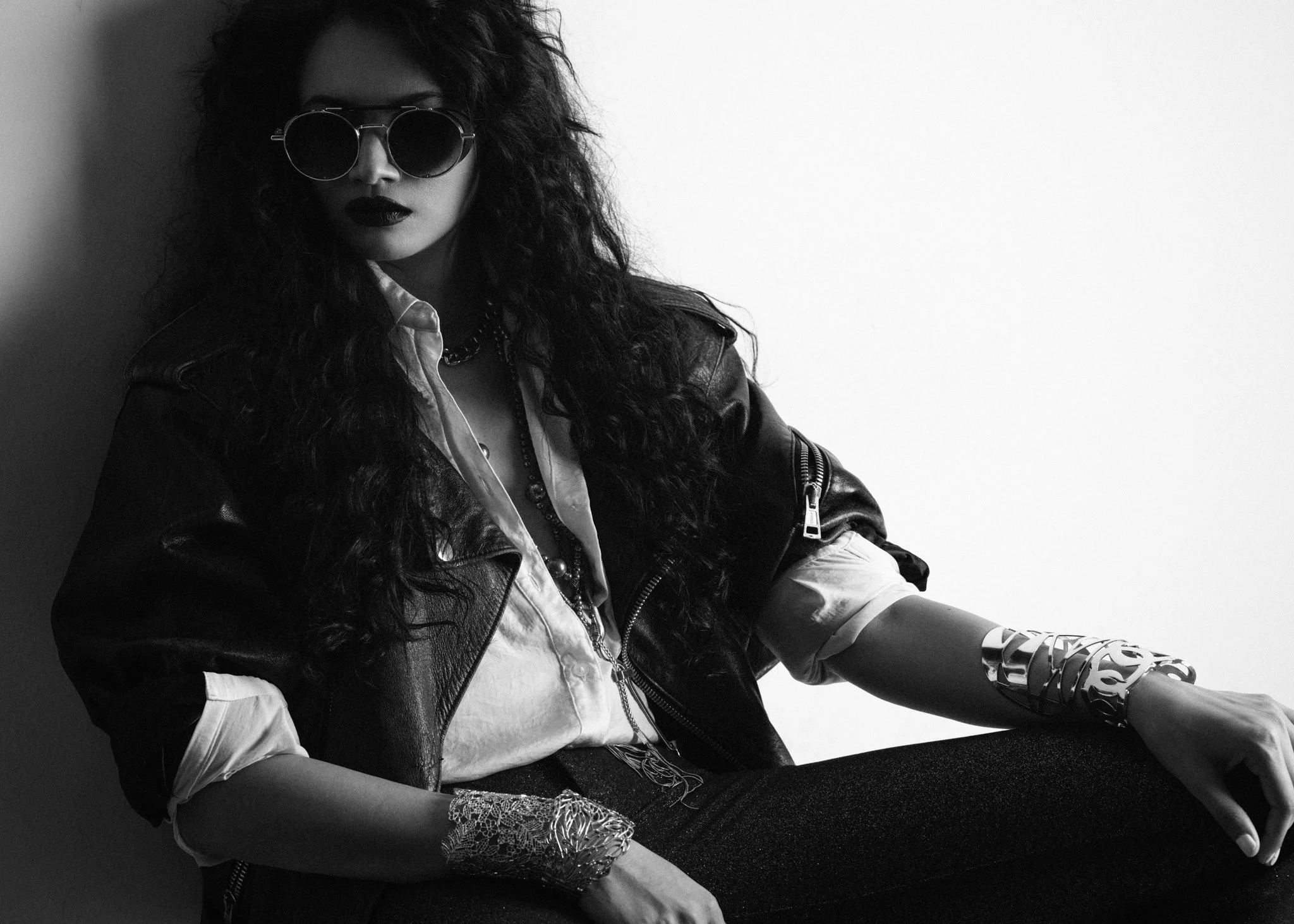 A woman with curly hair wearing sunglasses, a leather jacket, a button-up shirt, and multiple bracelets, sitting against a plain background.