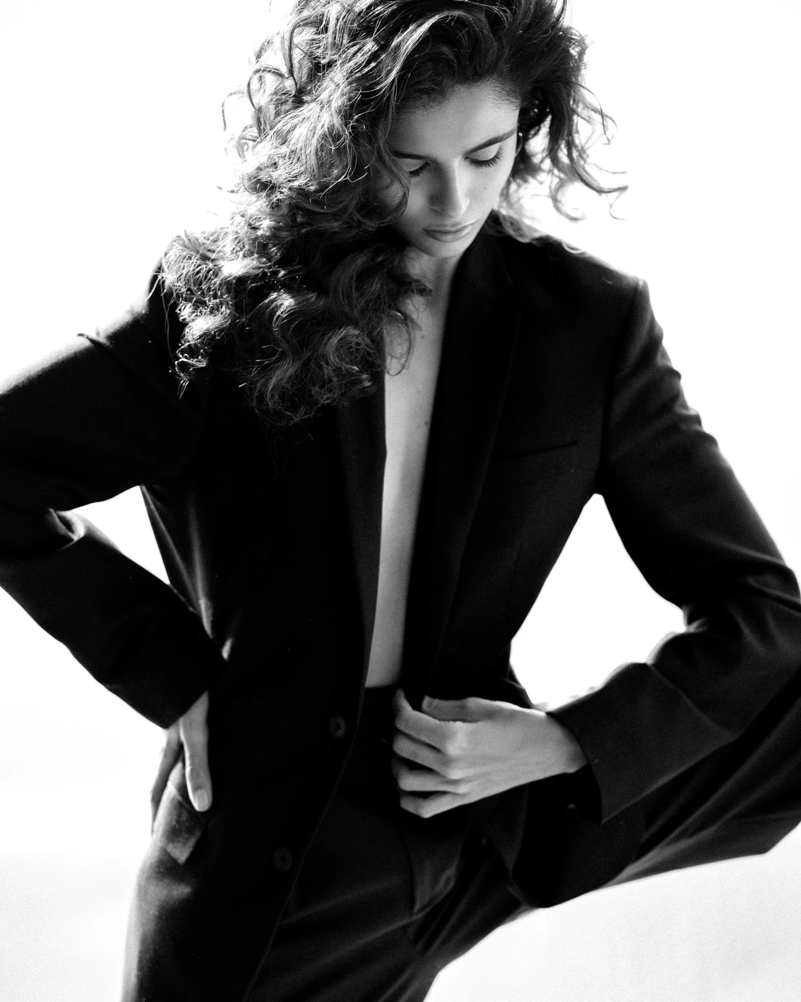 A black-and-white photograph of a woman with curly hair, dressed in a blazer and suit pants, posed with her head slightly bowed and hands on her hips.