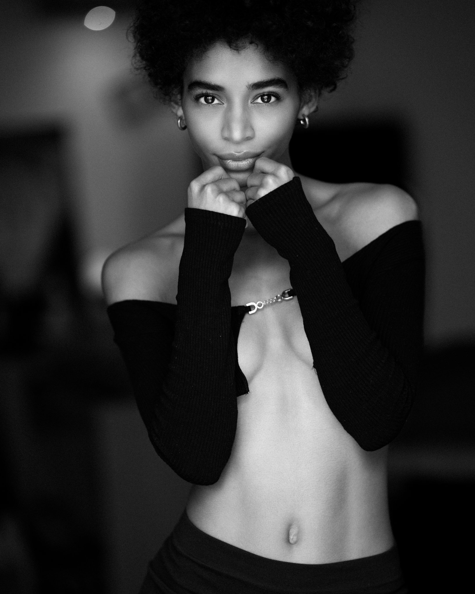 Black and white portrait of a young woman with curly hair, wearing off-the-shoulder long sleeves and jewelry, looking directly at the camera.