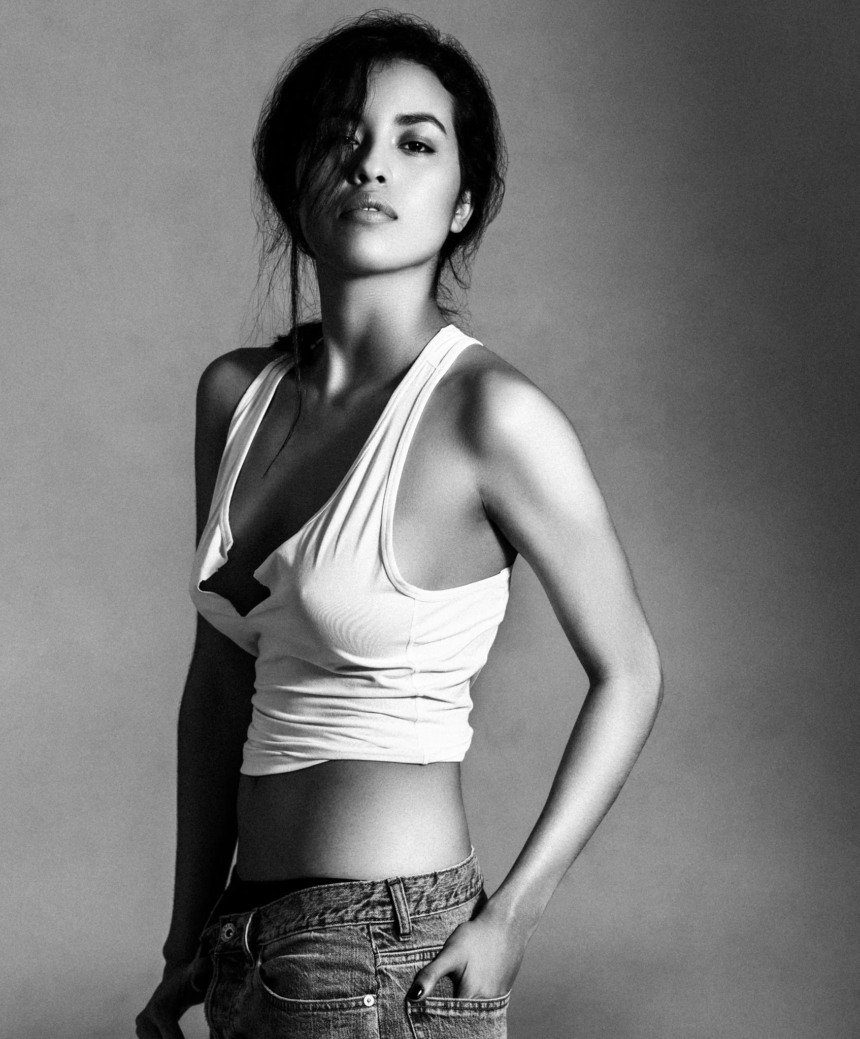 A black-and-white portrait of a woman with short, messy hair, wearing a tank top and jeans, against a plain background.