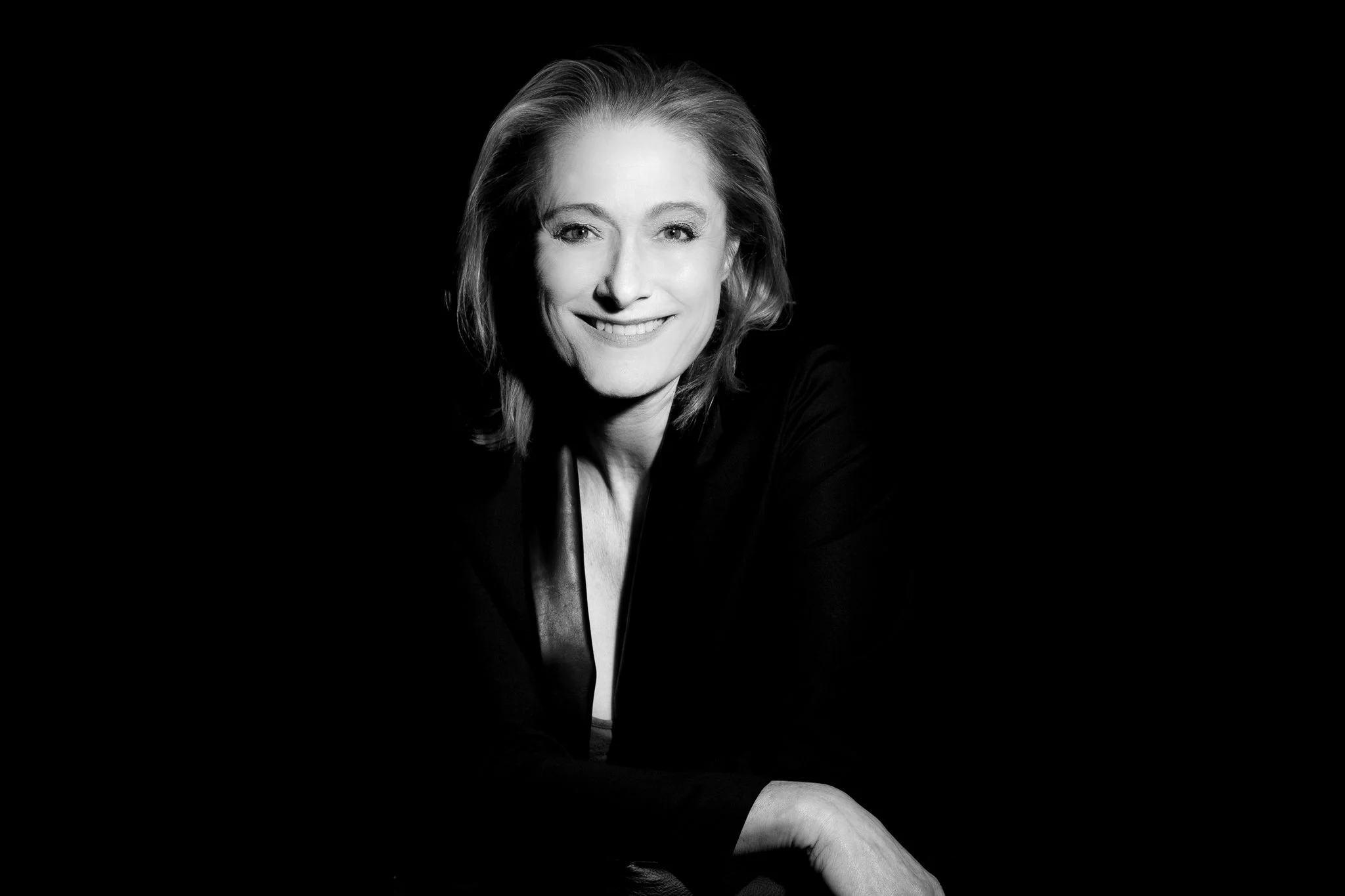 Black and white portrait of a smiling woman with wavy shoulder-length hair, wearing a dark blazer, against a dark background.