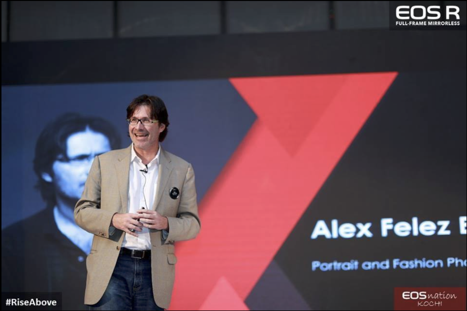 Alex F Buchholz speaking at a presentation, with a large black and red screen behind him displaying the text 'Alex Felez' and 'Portrait and Fashion Photography', and a partially visible person’s face on the screen.