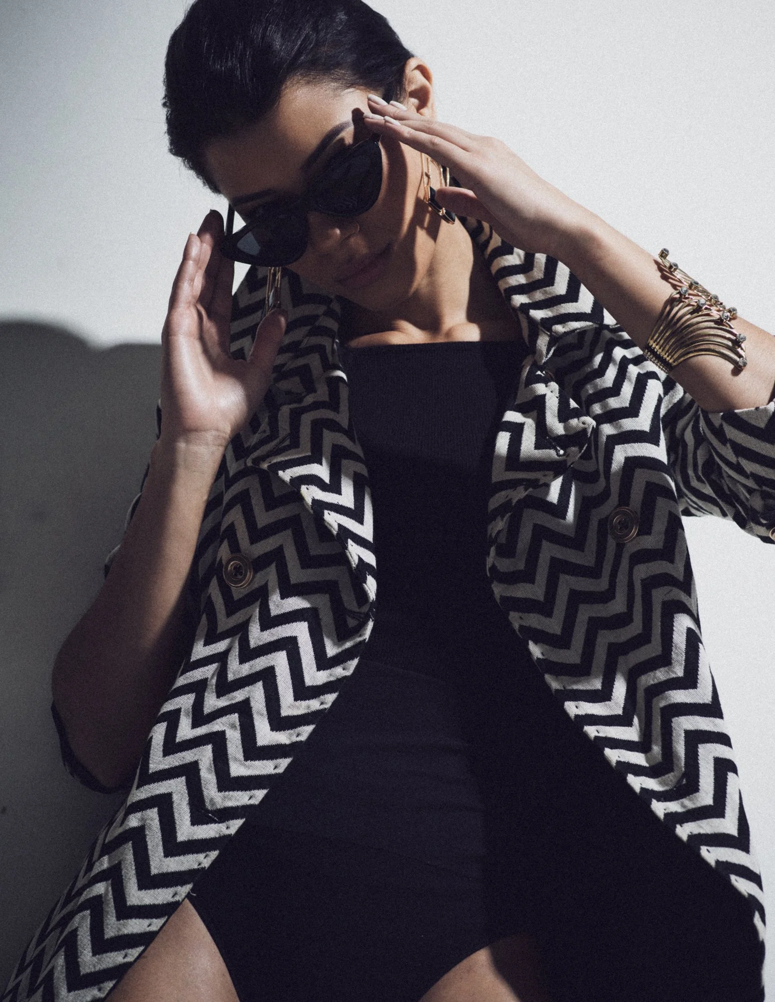 A woman with short dark hair wearing black sunglasses, a black dress, and a black and white chevron-patterned coat, poses against a plain background.