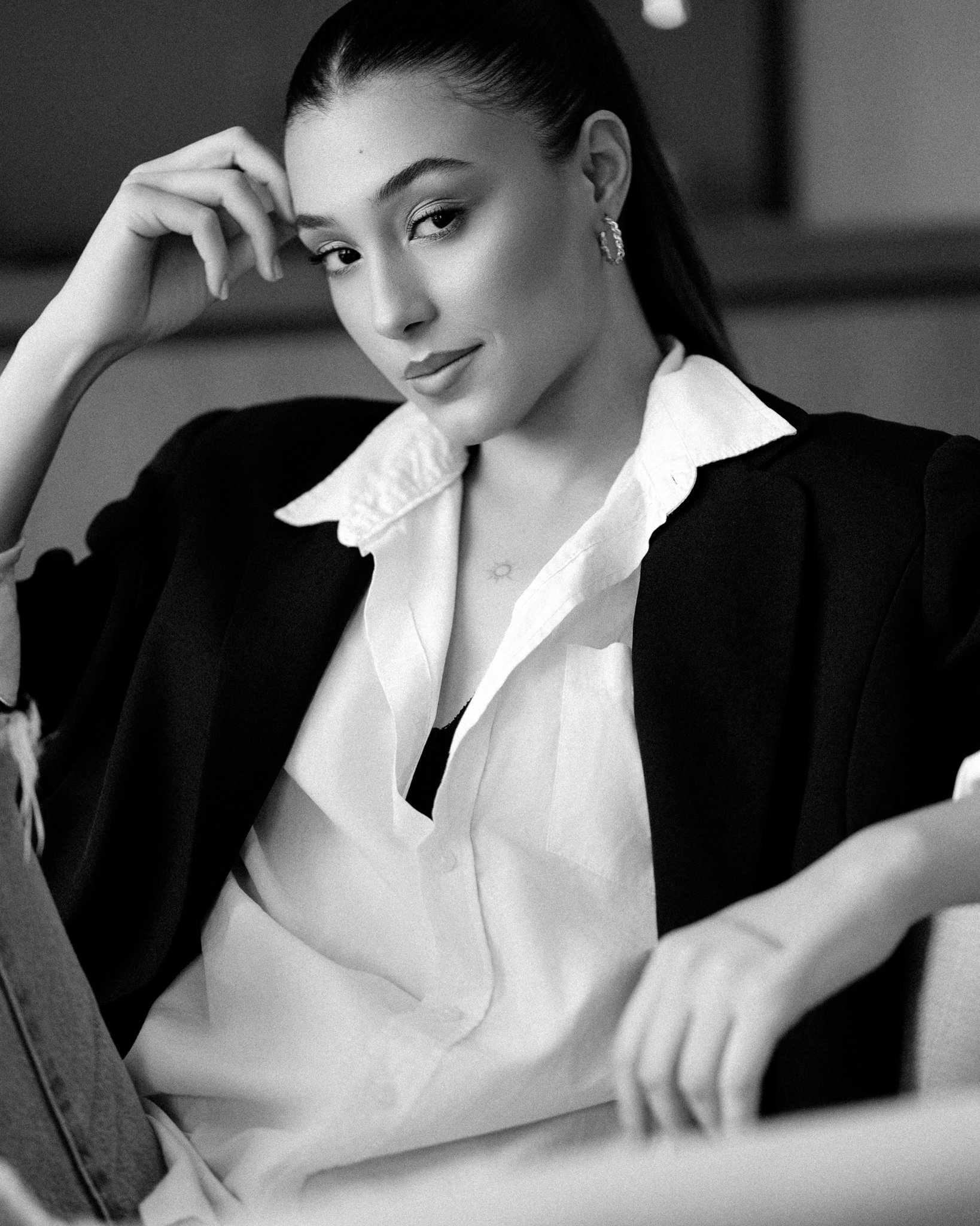 Black and white photo of a woman with dark hair pulled back, smiling slightly, wearing earrings, a white collared shirt under a dark jacket, sitting with one hand near her temple and the other resting on her lap.
