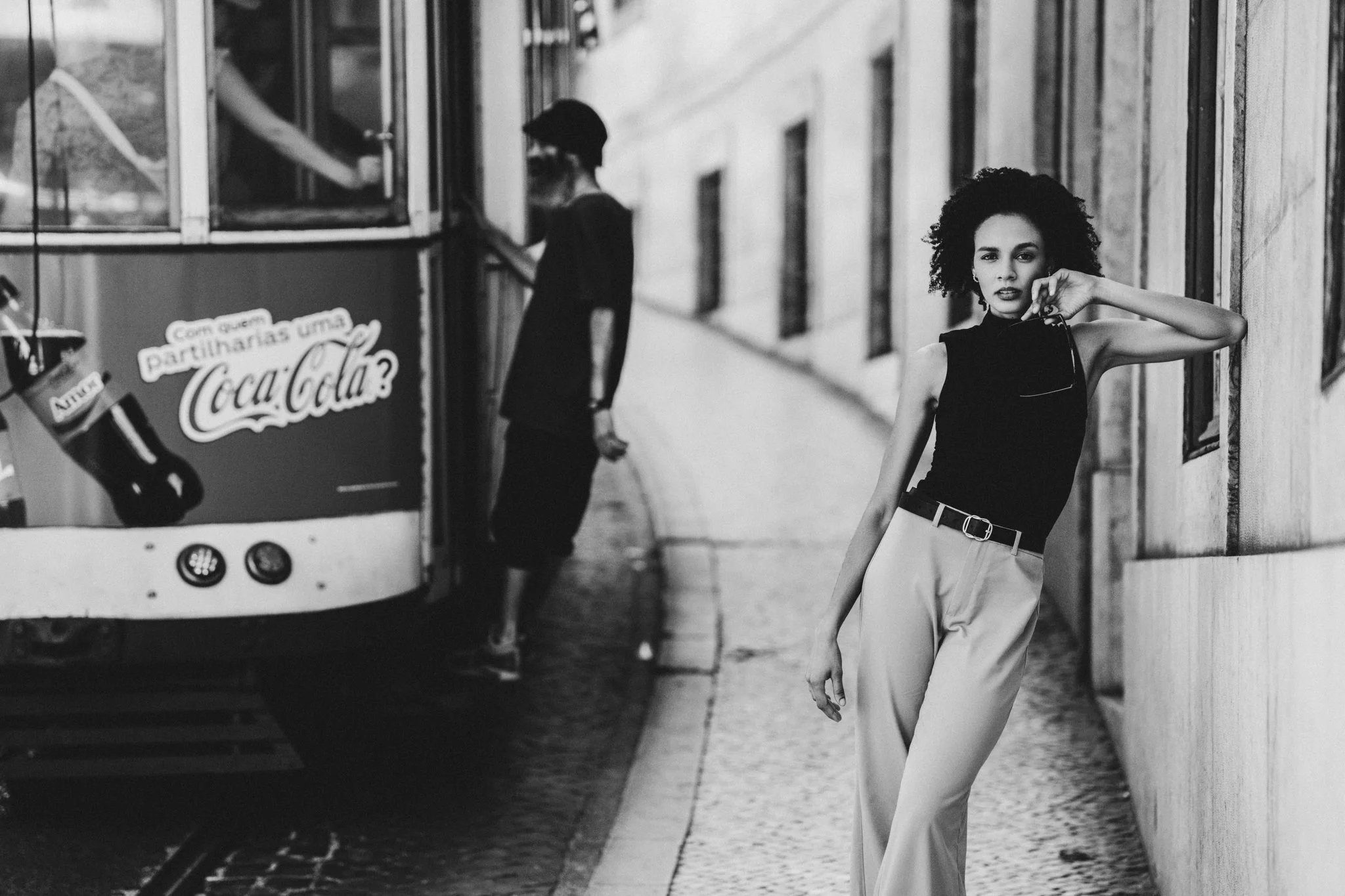A woman standing on a sidewalk leaning against a building, with a woman in back near a Coca-Cola vending machine. The image is black and white.