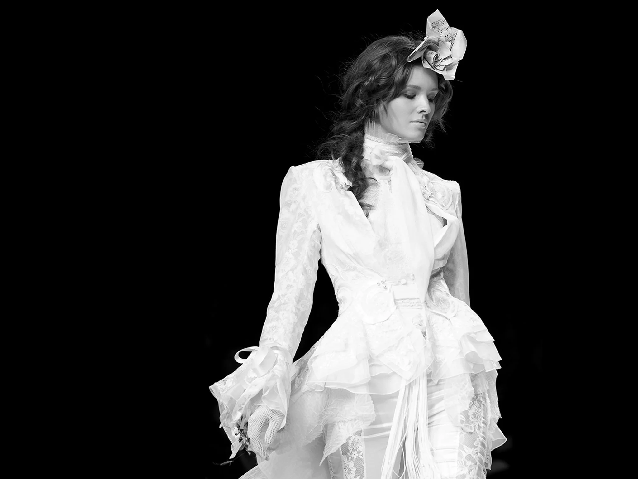 Black and white photo of a woman in a vintage wedding dress with lace details, a high collar, and puffed sleeves, wearing a large decorative bow and paper flower headpiece, looking down with her eyes closed.
