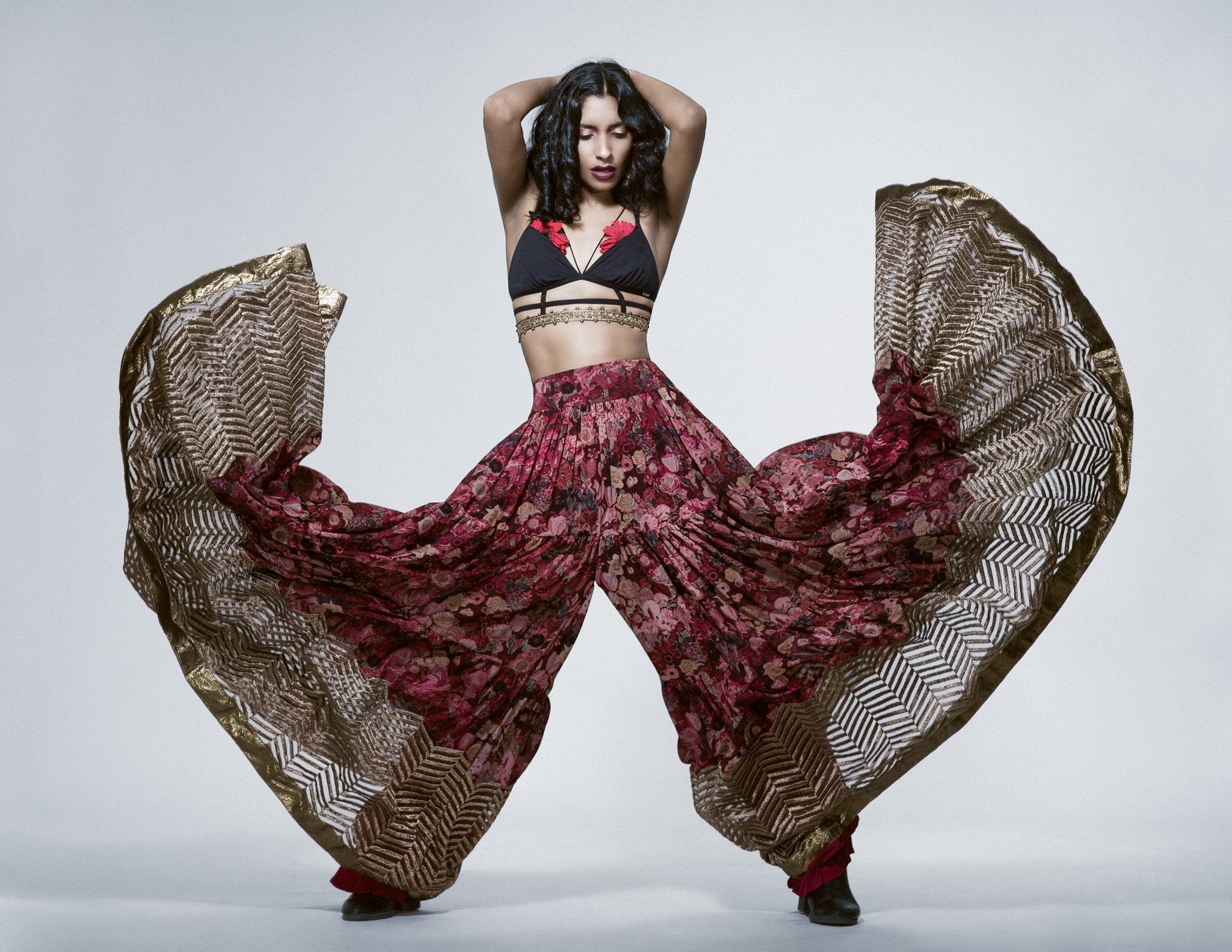 A woman standing with arms raised above her head, wearing a black and red crop top, a flowy skirt with floral pattern and dark footwear, surrounded by large patterned fabric pieces.