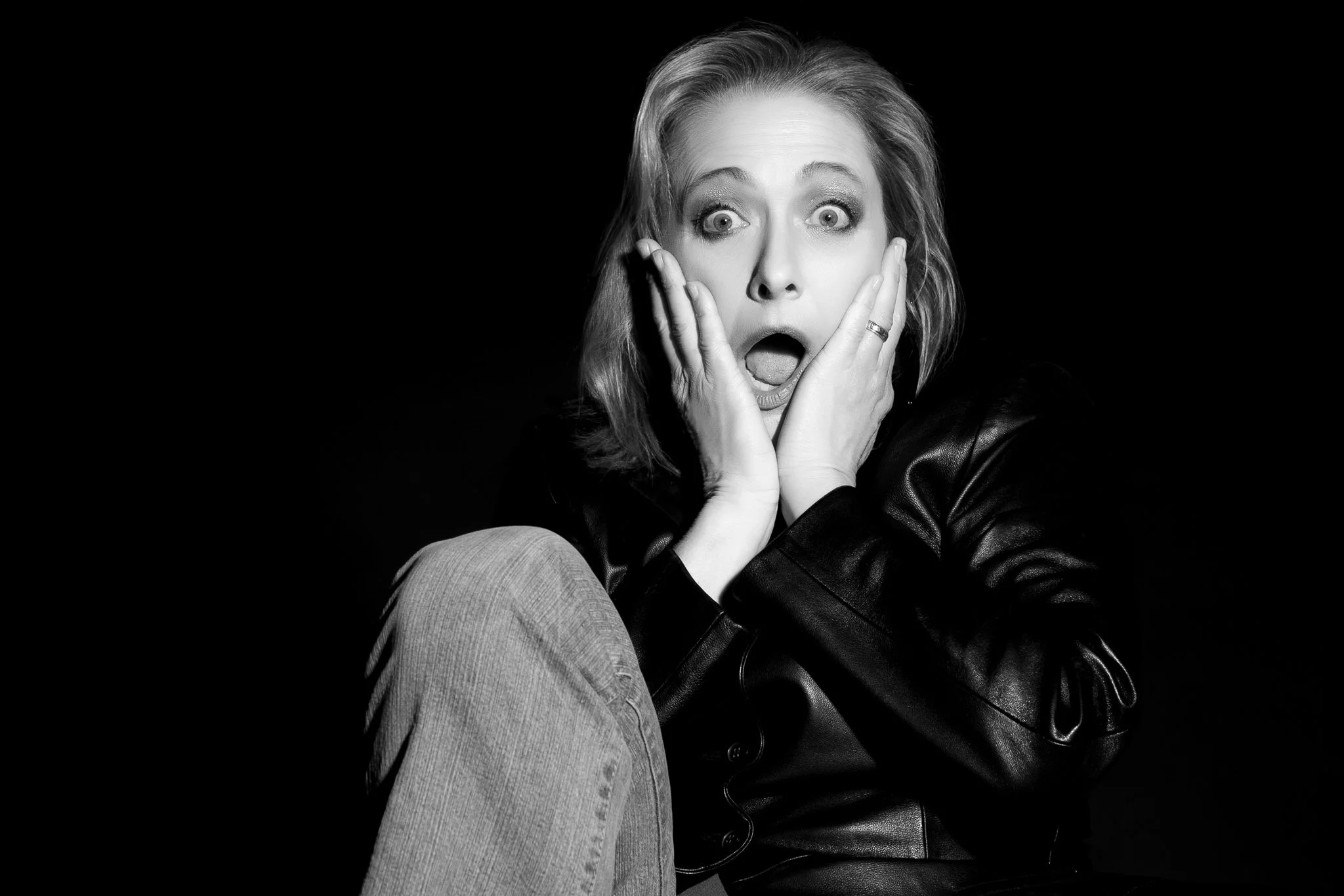 Black and white photo of a woman with an expression of shock, her eyes wide open, mouth agape, hands on her cheeks, wearing a leather jacket, with a dark background.