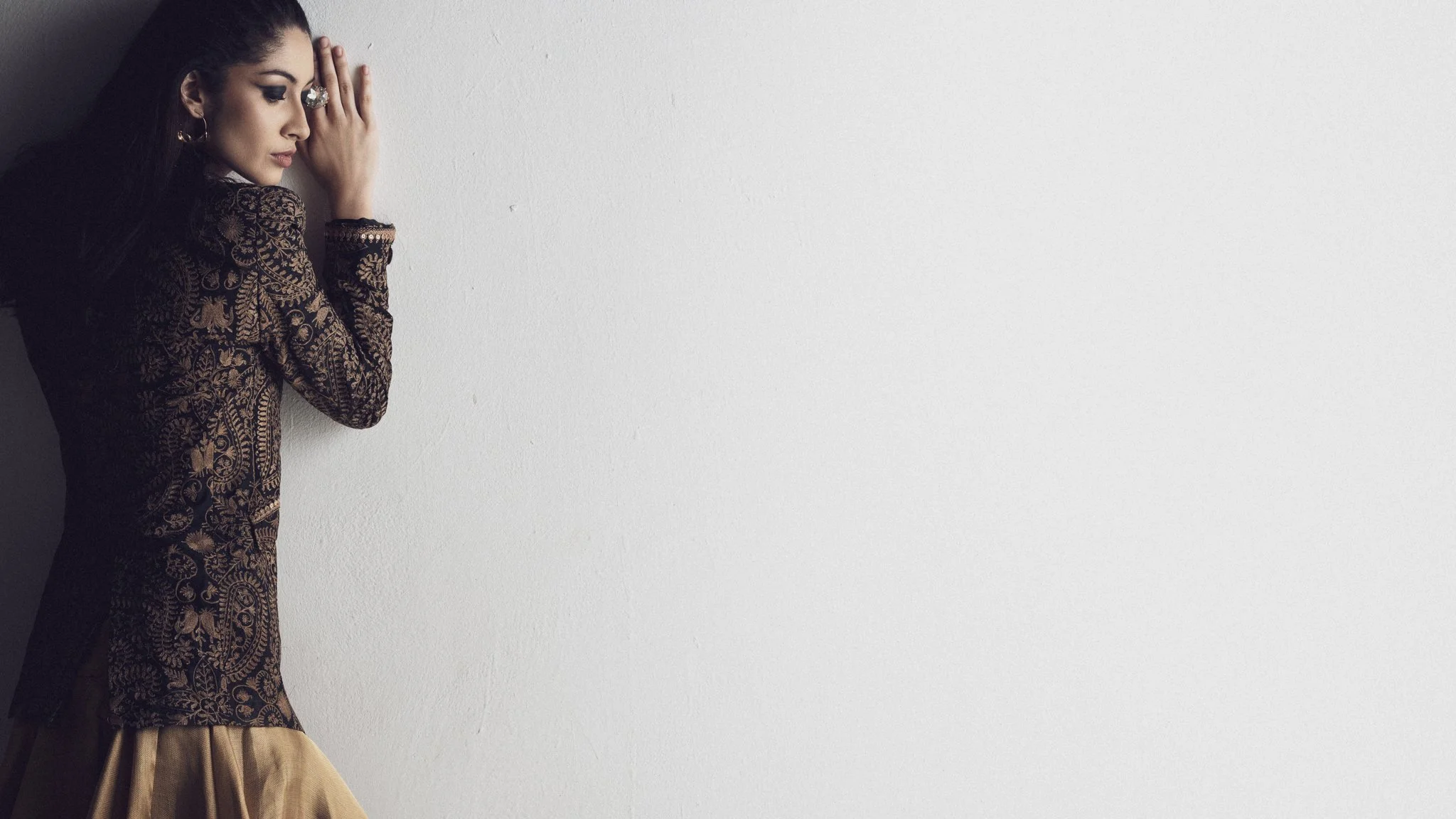 A woman with dark hair and makeup, wearing a patterned dress, is standing against a plain white wall, with her hands pressed together near her face in a prayer-like pose.