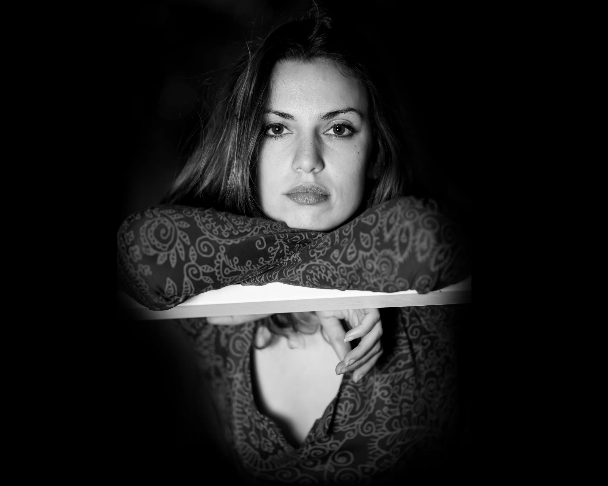 Black and white portrait of a woman with wavy hair, resting her chin on her folded arms, wearing a patterned top with a cutout, looking directly at the camera. The background is dark, emphasizing her face and expression.