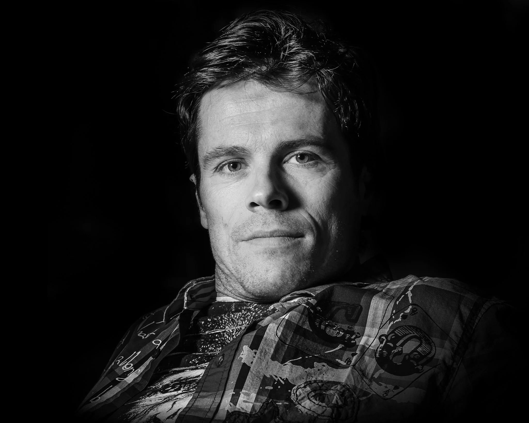 Black and white portrait of a man with wavy hair, gazing directly at the camera, with a subtle smile, wearing a patterned shirt.