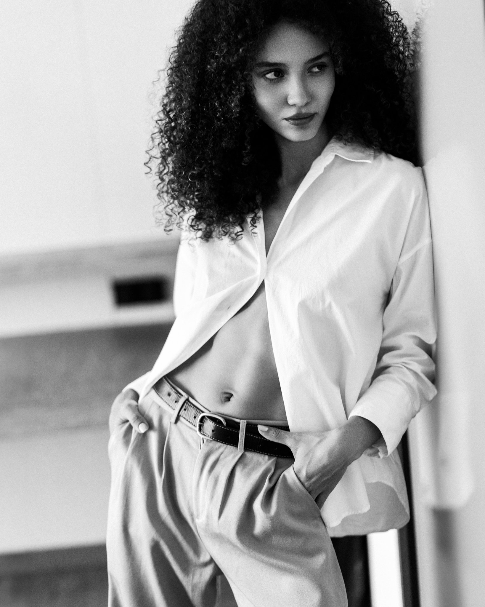 A young woman with curly hair leans against a wall, wearing an open white shirt, beige trousers, and a belt, with her hand in her pocket, looking sideways in a black-and-white photo.