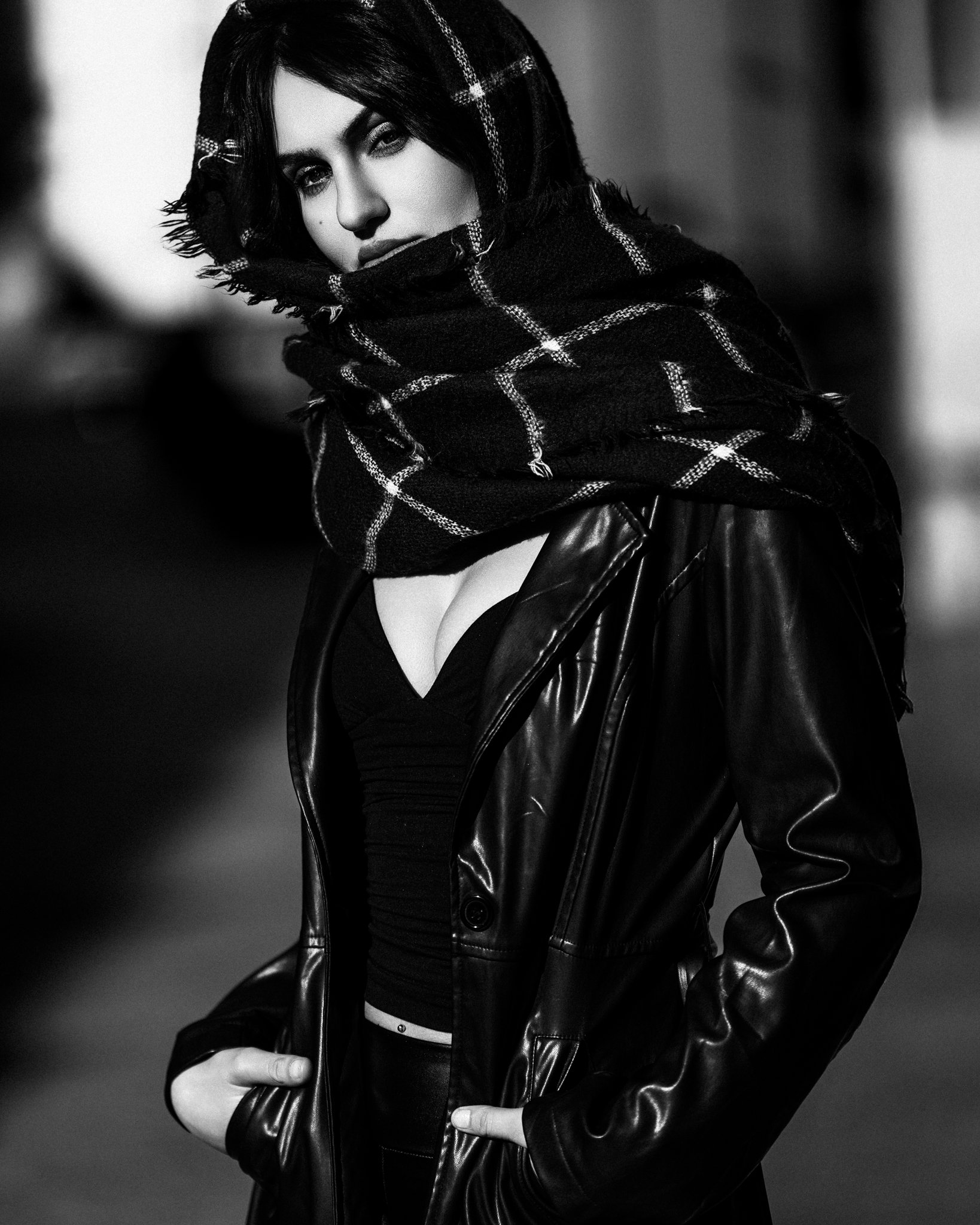 Black and white photo of a woman with dark hair, wearing a leather jacket and a plaid scarf, looking at the camera with a serious expression.