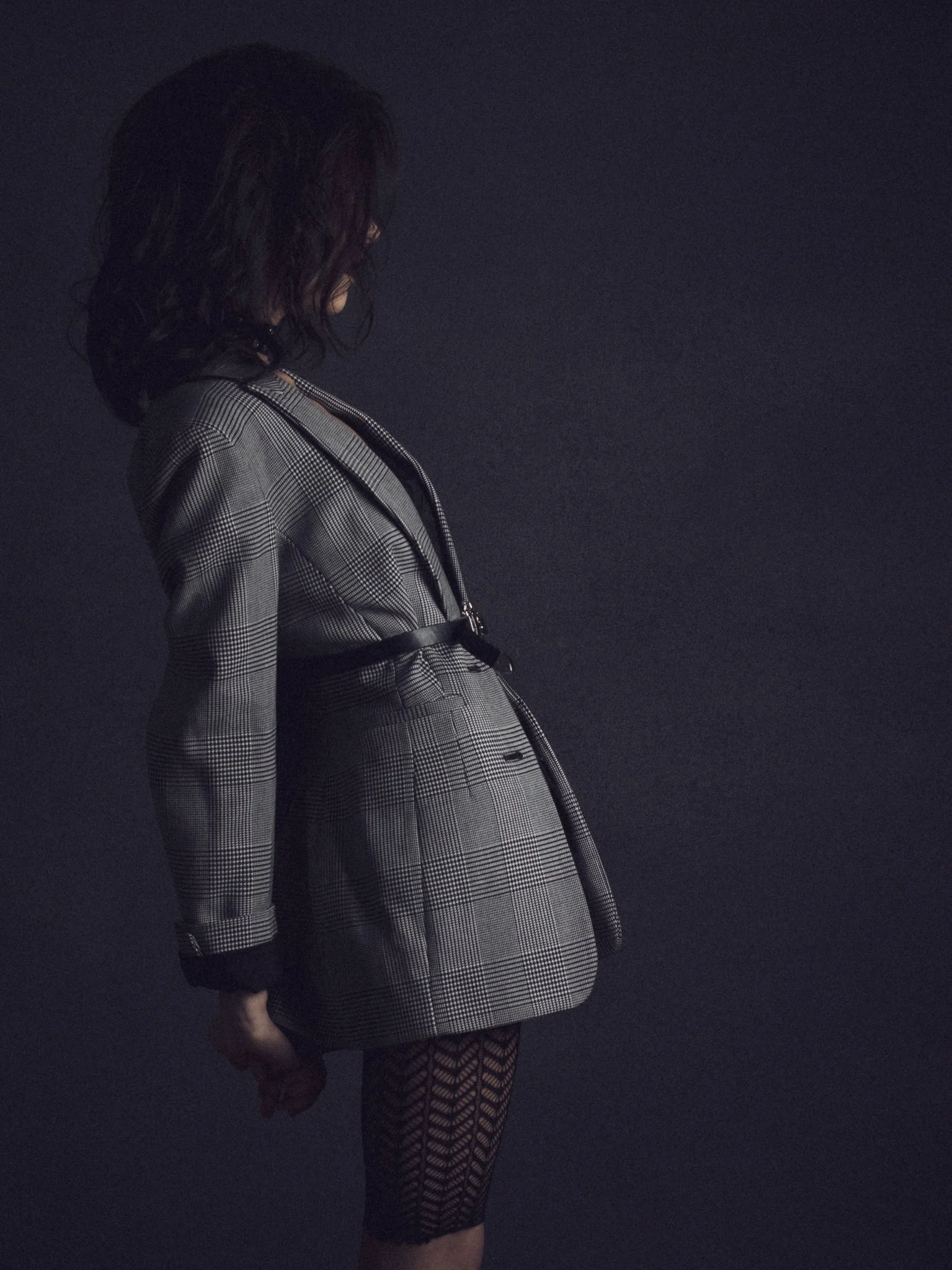 A woman with dark, wavy hair wearing a plaid blazer with matching skirt, sheer patterned tights, and black shoes, standing against a dark background.
