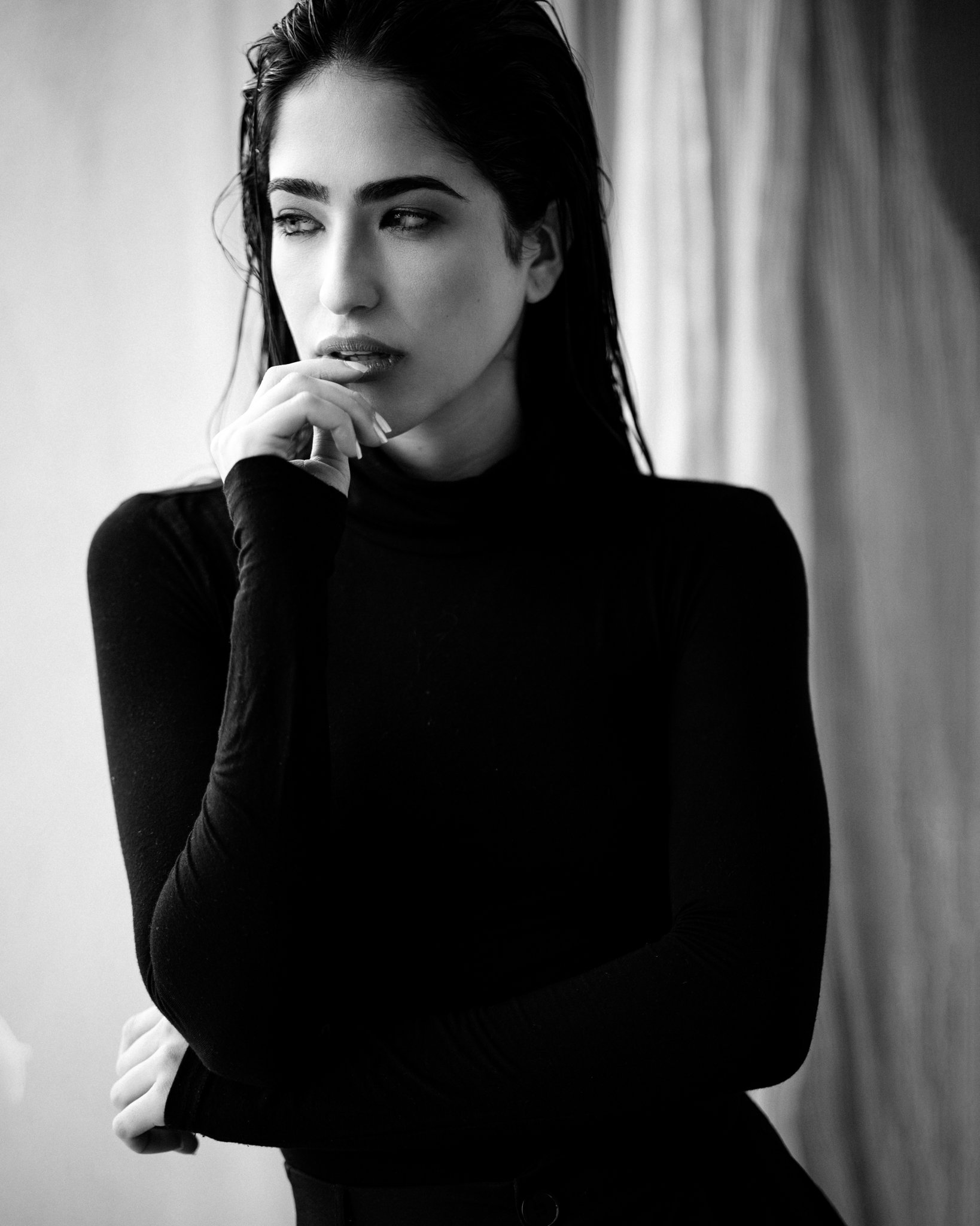 A black and white photo of a woman with dark hair wearing a black turtleneck. She has a thoughtful expression, with one hand touching her lips and her gaze directed sideways.