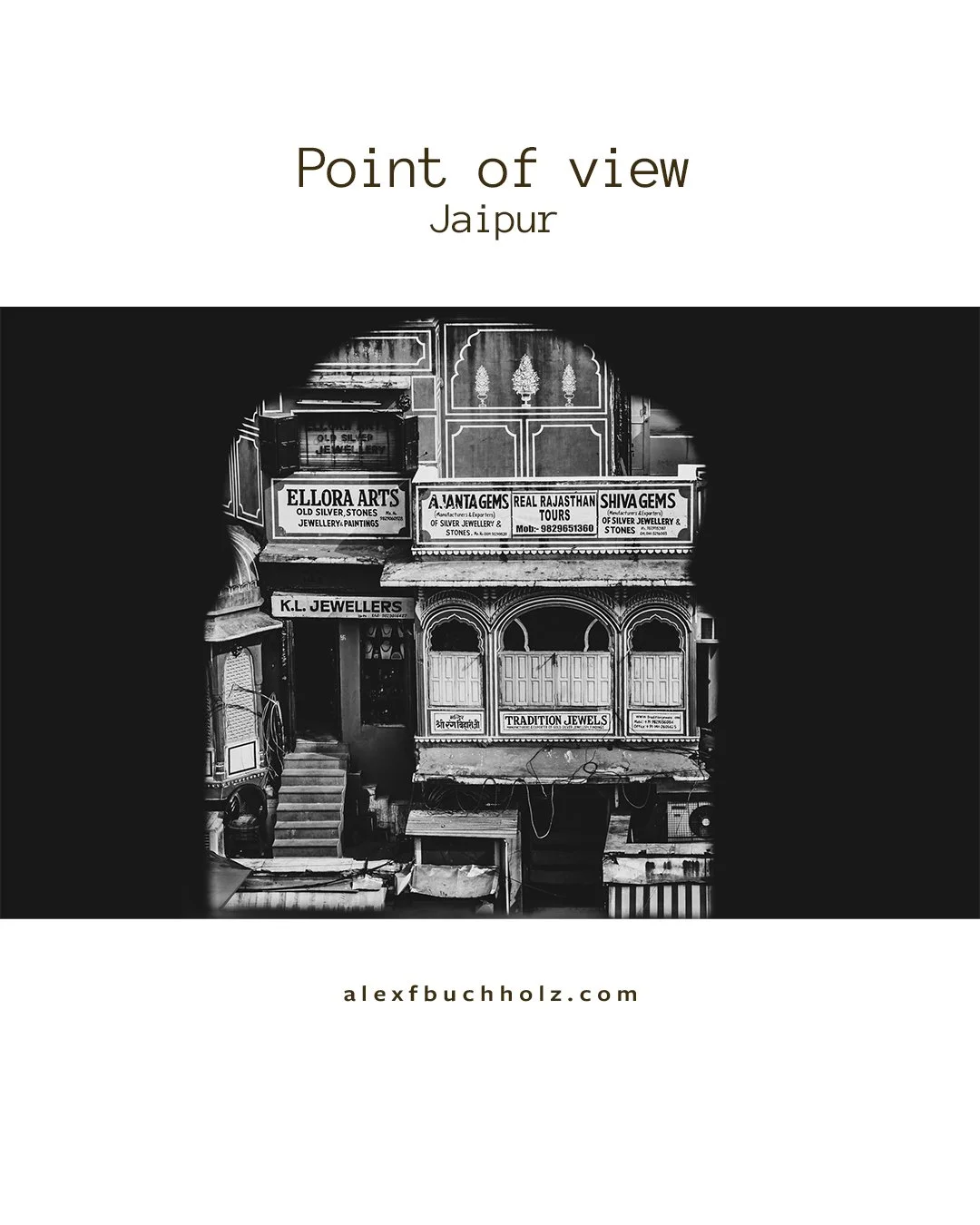 Black and white photograph of a street view in Jaipur, India, featuring two adjacent buildings with signs advertising jewelry stores and tours, viewed through a circular opening.