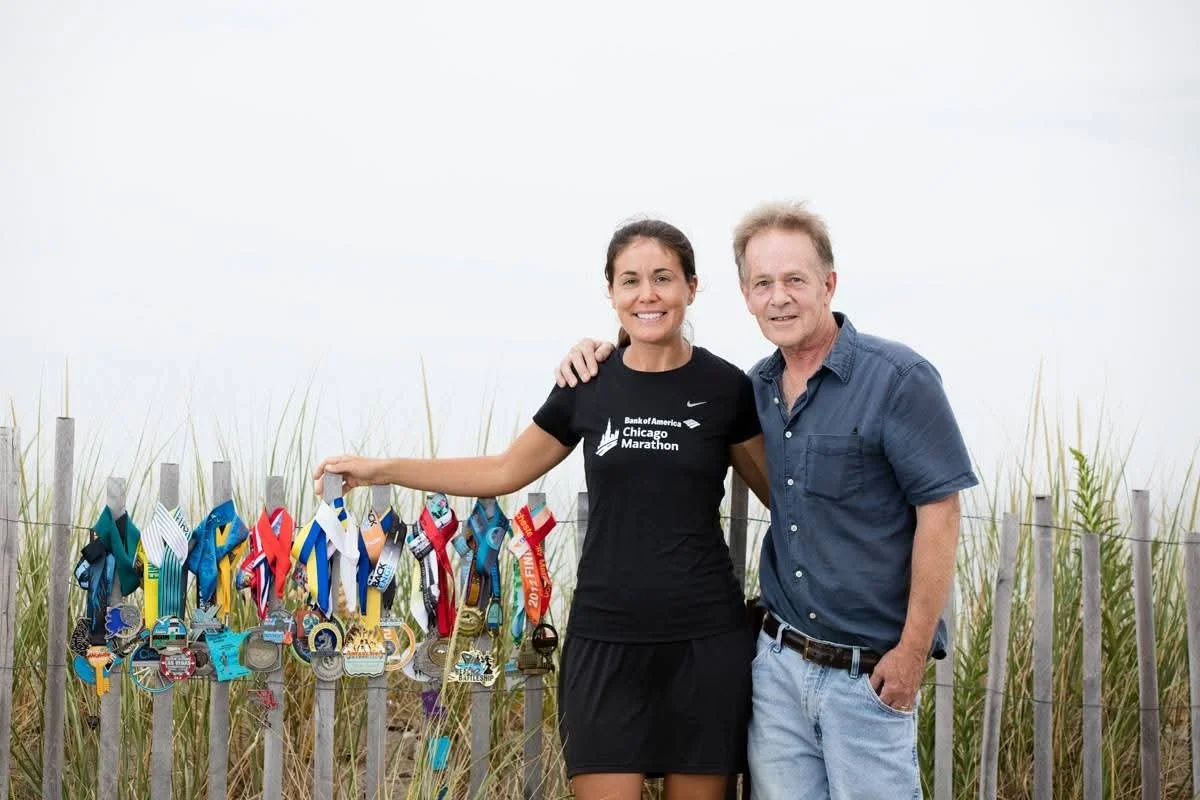 Woman wearing a Chicago Marathon shirt poses with her dad and her marathon medals draped over a fence