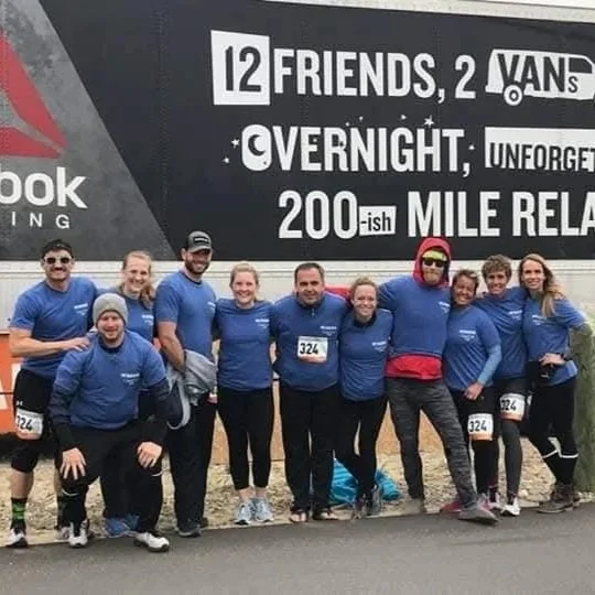 Group of eleven people wearing matching blue shirts in front of a banner for a relay race