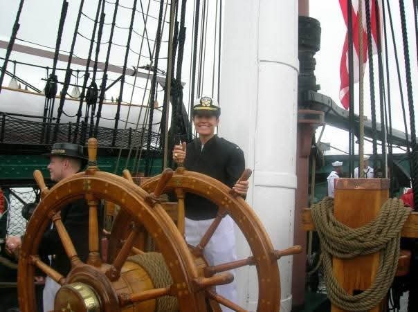 Woman wearing U.S. Navy uniform stands behind the wheel of a ship